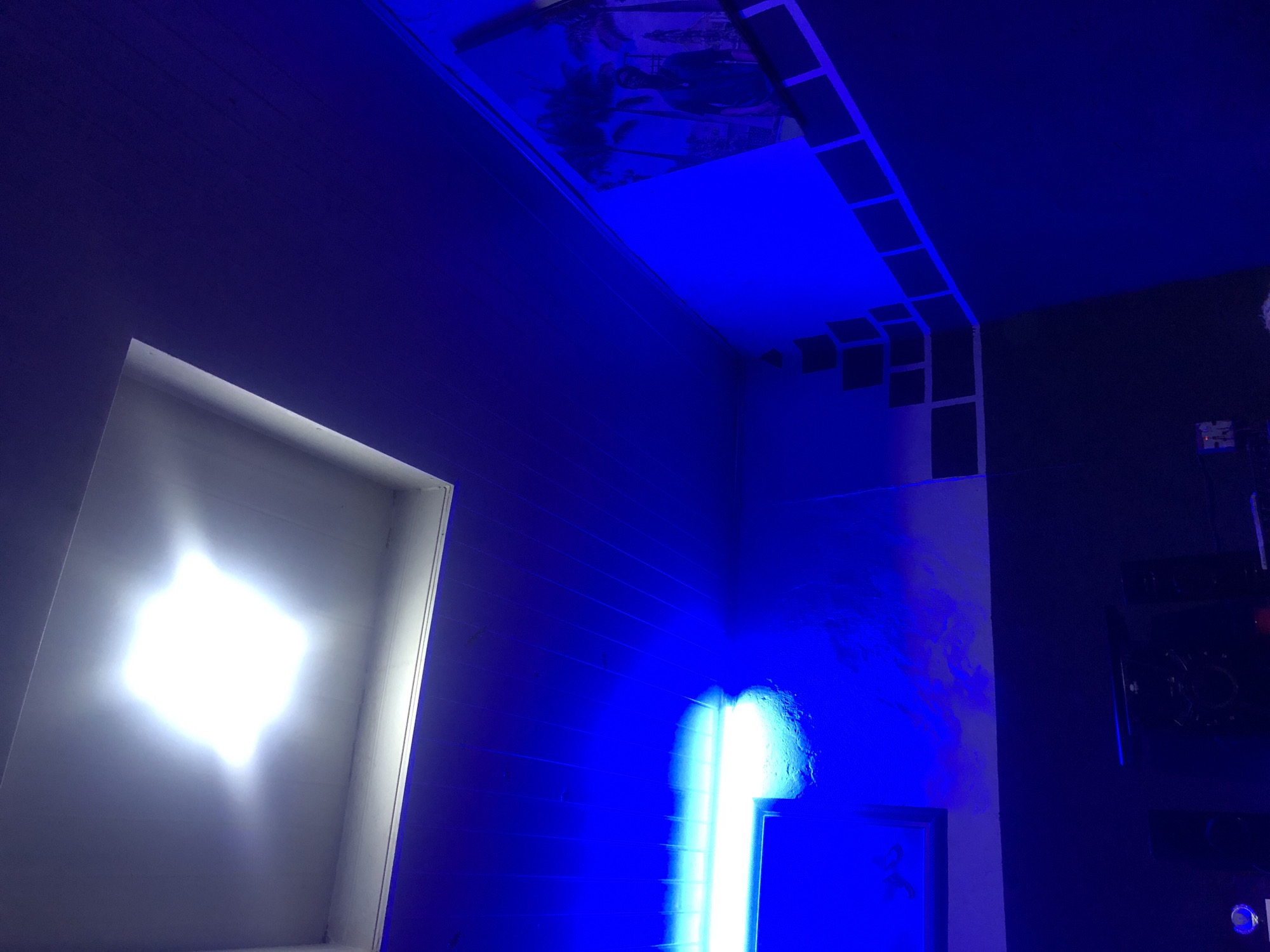 The image displays an interior room in the city of Unknown, Unknown, characterized by dual lighting sources: a central, bright white overhead light and multiple blue lights. The ceiling is primarily dark blue, with a square, recessed white fixture housing the overhead white light in the upper-left quadrant. A linear, horizontal fluorescent light fixture emitting a bright blue light is mounted on the upper-left wall. Below this blue light, the wall surface exhibits irregular texture. The upper sections of the visible walls are uniformly illuminated in a vibrant blue hue. The lower sections of the walls are painted a dark, non-reflective color, forming a clear horizontal demarcation line. On the upper-right wall, a framed image or poster depicts palm tree silhouettes and a dark human figure against a blue background, consistent with the room's lighting. Below this poster and extending along the dark wall section are multiple square and rectangular dark panels, arranged in both diagonal and horizontal patterns. In the lower-left foreground, two dark audio speakers are visible, one a central unit and another a tower speaker. A small, light-colored object with a reflective surface is present in the extreme lower-left. The overall illumination creates strong blue color casts across the surfaces.