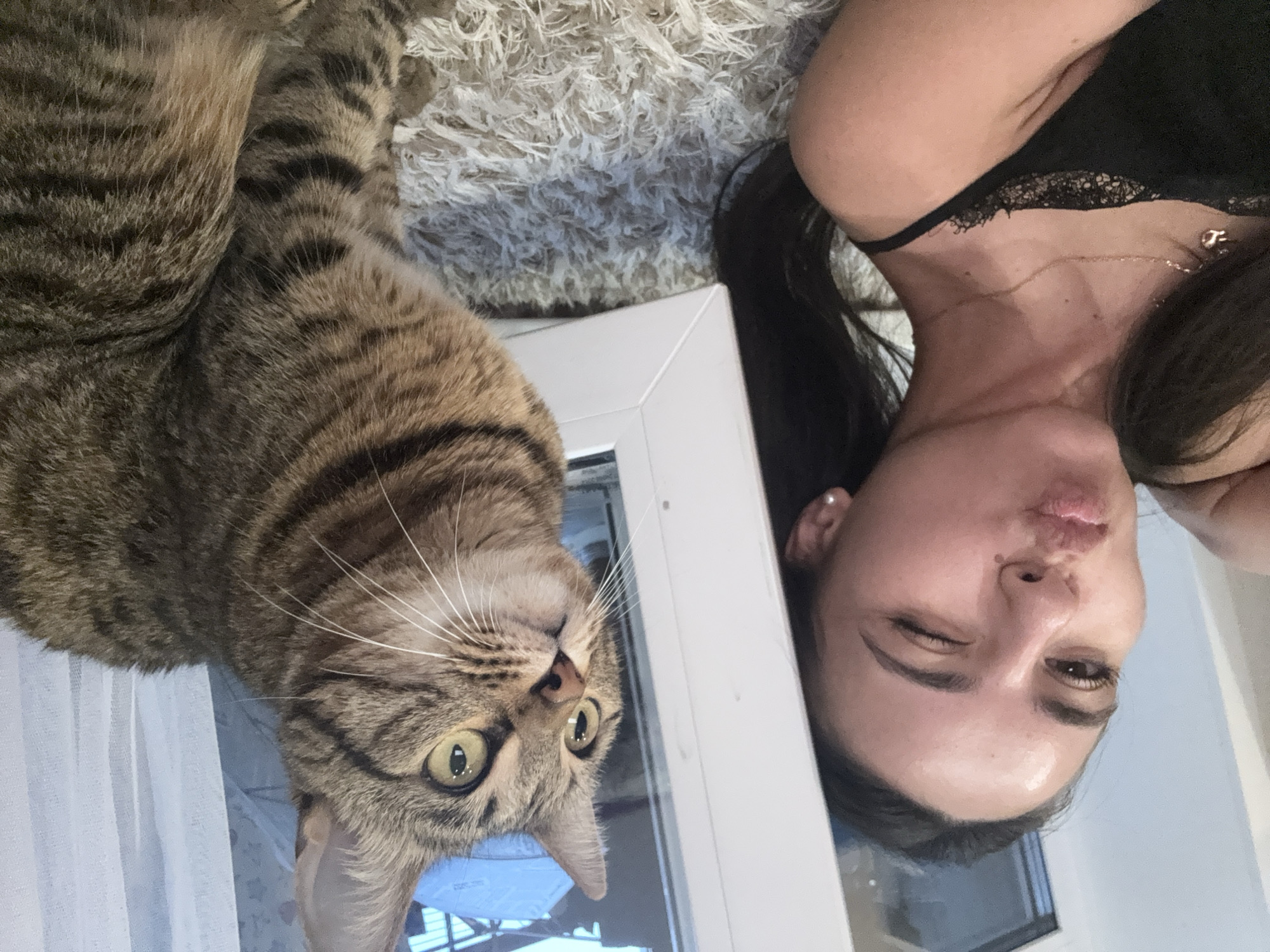 A young woman is lying on a fluffy rug, puckering her lips and winking at the camera. Beside her, a tabby cat sits upright, looking to the right with bright yellow eyes. The woman is wearing a black lace-trimmed top, and a delicate necklace with a charm. The background appears to be an indoor setting with a large window and sheer curtains.