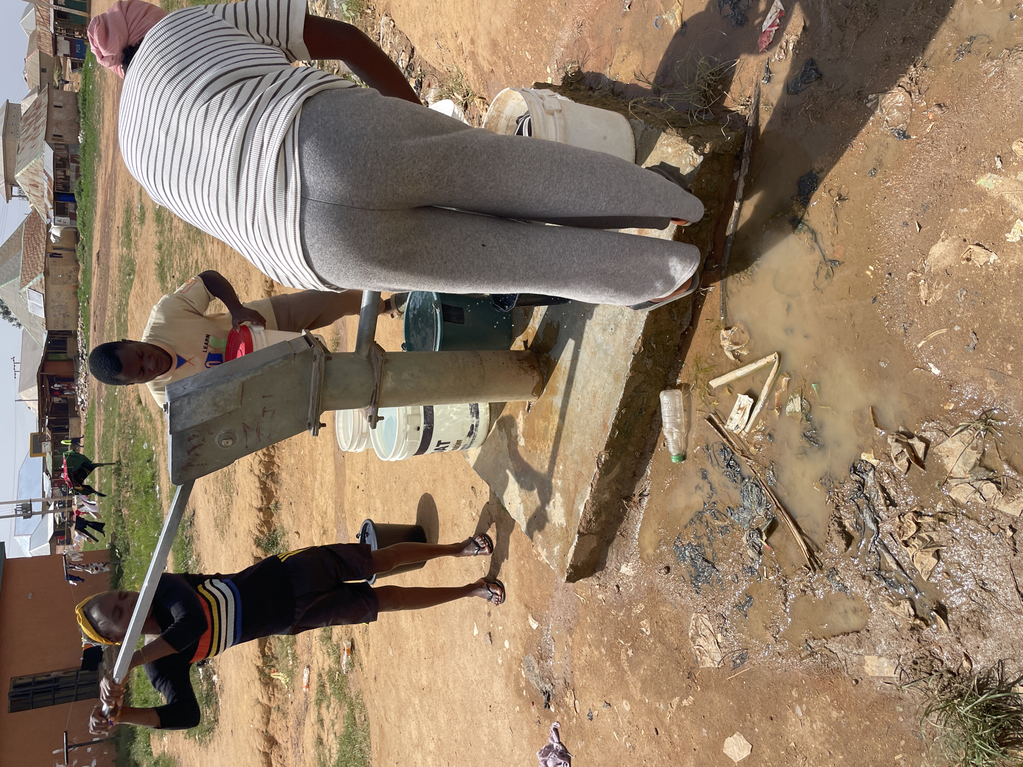 The image depicts a scene in Durimi, Nigeria, where three individuals are interacting with a water pump. A woman with a pink head covering and a striped shirt stands on the pump's concrete base. A man in a tan shirt is filling a white bucket from the pump. A young woman in a dark shirt with colorful stripes is operating the pump handle. Several white buckets are stacked near the pump. The ground around the pump is muddy and littered with trash, including a plastic bottle. Buildings are visible in the background.