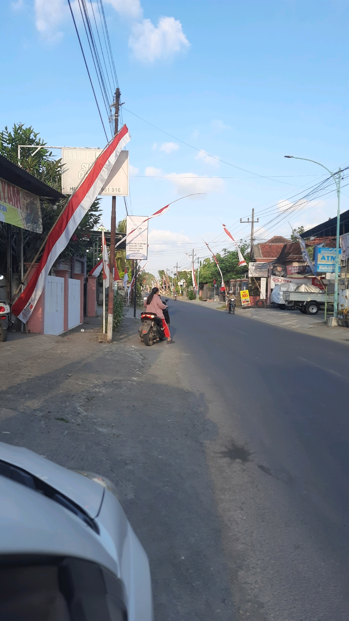 This is a full shot of a street in Bangorejo, Indonesia, featuring a clear blue sky with scattered clouds. The street is lined with buildings on both sides, displaying a mix of residential and commercial structures. Red and white Indonesian flags, indicating a celebration or national holiday, are strung across the street and attached to poles. A woman is riding a motorcycle on the road, wearing a pink long-sleeved shirt and red pants. She is accompanied by another person on the back of the motorcycle. In the foreground, a portion of a white car is visible, suggesting the vantage point of the photographer. The street itself is paved and appears to be in moderate condition, with some minor wear and tear visible on the asphalt. There are trees and vegetation along the sides of the street, adding to the natural environment of the scene. Several signs are mounted on poles, some of which are advertising businesses or services, like an ATM. The overall impression is a peaceful, everyday scene in a small Indonesian town, with a festive atmosphere due to the presence of the flags.