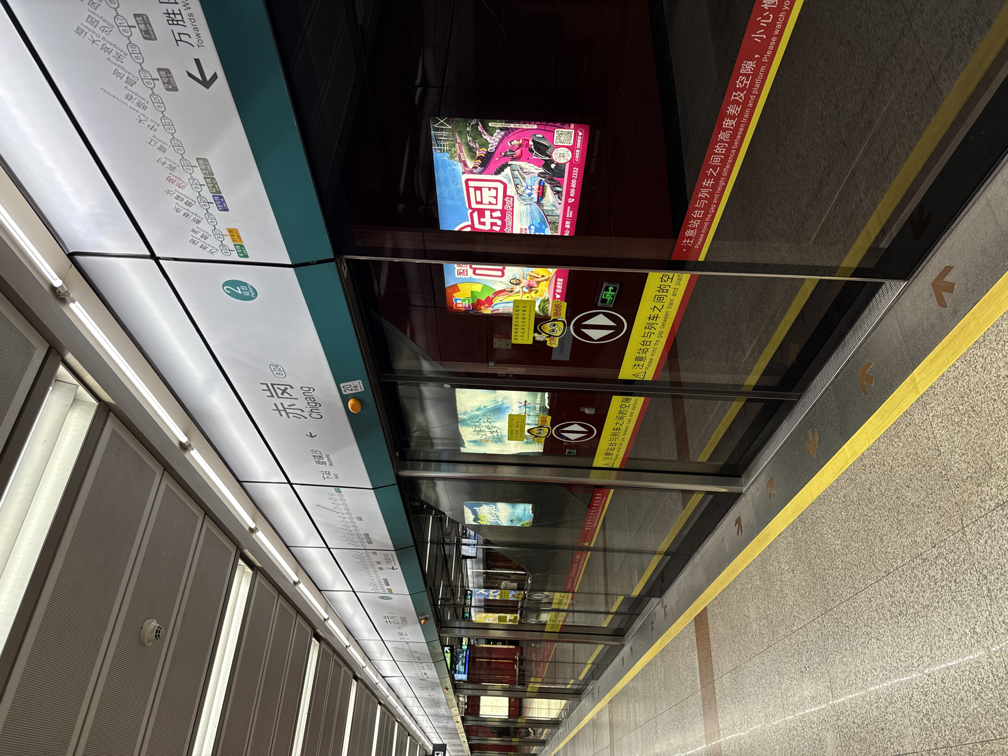 This is a photograph taken on a subway platform in Haizhu District, China, likely during daylight hours as indicated by the bright interior lighting. The perspective is from inside a subway car, looking out through the doors onto the platform.
The main subjects are the subway platform itself and the glass doors of the train. On the platform, there's a digital display showing a subway map, with station names like "Chigang," "Mobedisha," and "Xiaogang" visible. An advertisement for an "Adventure Park" is displayed on a screen above the platform doors. Yellow and brown lines on the floor indicate the safety zone and boarding areas. The doors of the subway car are large and reflective, showing the interior of the train and the platform scenery.
The scene is a typical urban subway environment, characterized by clean, modern infrastructure. There are no people clearly visible in the photograph, but the setup suggests an active transit setting. The text visible includes station names in both Chinese and English, as well as directional arrows and safety warnings in Chinese and English, such as "Please mind the gap between train and platform." The overall impression is one of order and functionality, common to public transportation systems. The photo seems to be taken from a slightly elevated angle, looking down and across the platform.