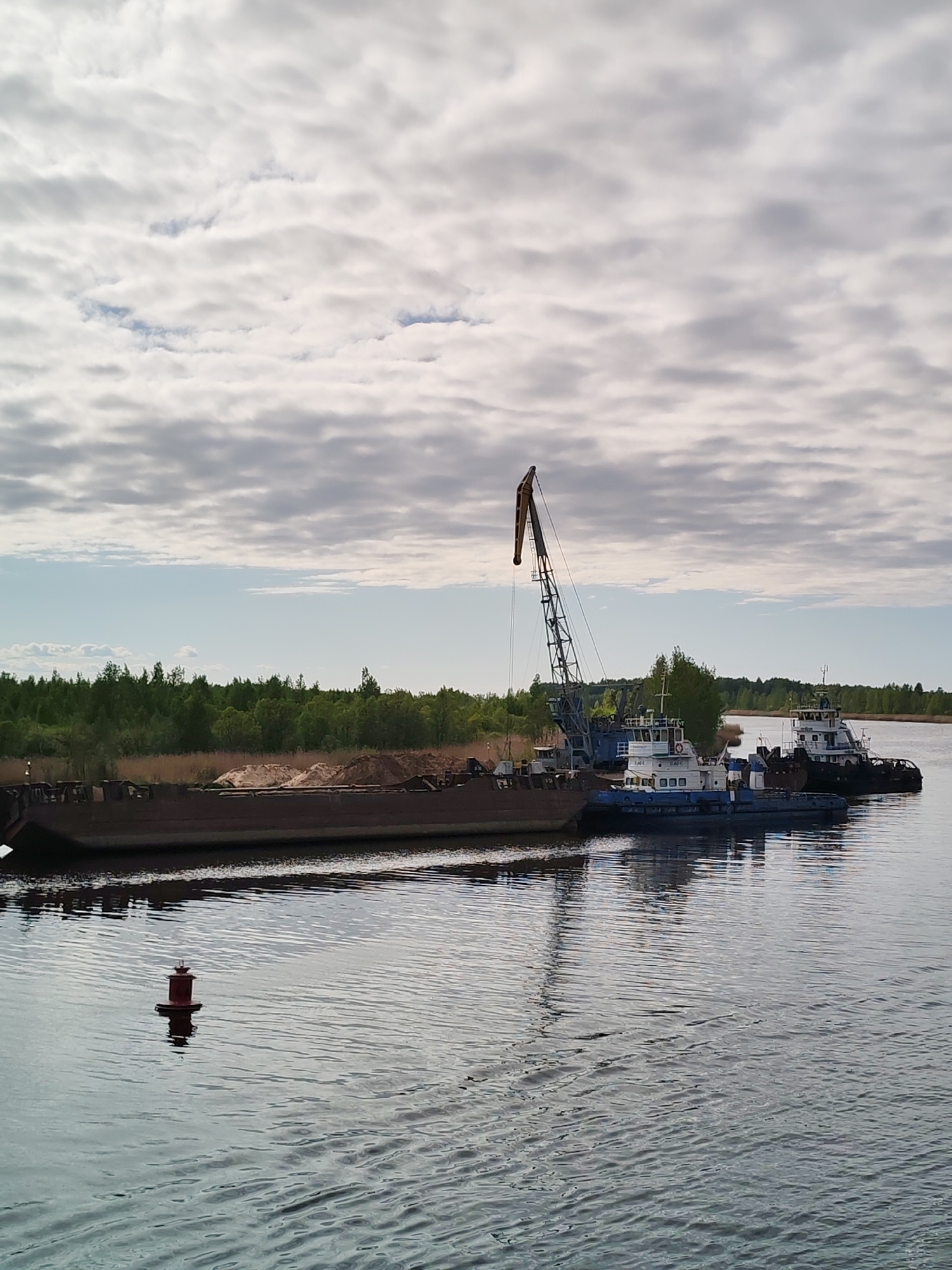 The image depicts a river scene at Ozernoe Ust'e, Russia. A long, dark barge filled with sand or gravel is docked alongside a blue and white tugboat with a large crane. A second, similar tugboat is positioned behind the first. A red buoy is visible in the foreground of the water, and a tree line stretches along the horizon. The sky is overcast with mottled clouds.