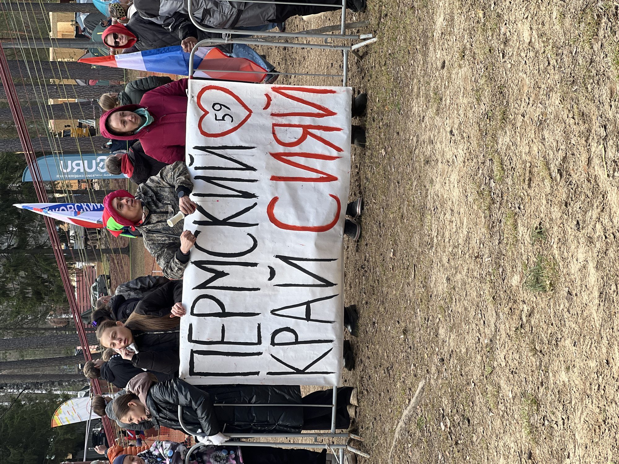 The image shows a group of people holding a sign that reads "Пермский Край Сияй" in Russian. The sign also has a heart with the number 59 inside it. The people are standing outdoors, in what appears to be a park or wooded area. The ground is covered in dirt and some grass. In the background, there are trees and a building. The sign "URU" is visible in the background. The image was likely taken in the city of Lesnoye, Russia.