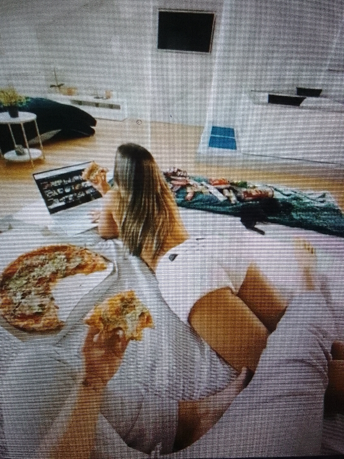 The image depicts an indoor scene within a modern residential space, characterized by light-colored walls and wooden flooring, located in Unknown, Unknown. A single individual is present, viewed from the back. This person has long, light brown hair and is lying prone on a light-colored duvet or blanket, facing a laptop with an active screen displaying multiple images. The individual is holding a slice of pizza to their mouth with their right hand. They are dressed in light-colored shorts. To the person's left, a partially consumed pizza is situated in a white cardboard box on the duvet. Another detached piece of pizza rests below the main box. In the background, a dark, rectangular television is mounted on the wall. A dark sofa and a two-tiered side table are visible in the upper left. A patterned dark green blanket is laid out on the floor to the right, adjacent to a small blue rectangular object affixed to the wall.