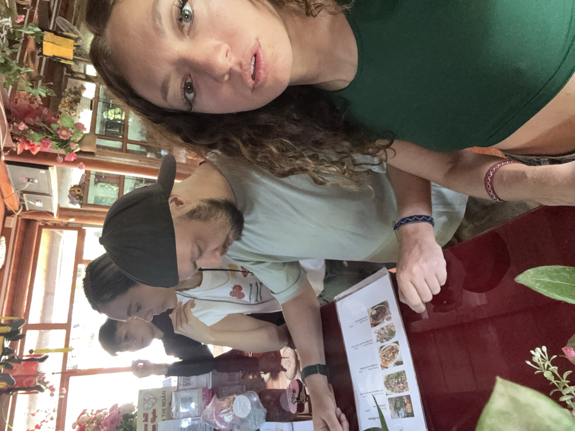The image shows a group of four people at a restaurant, potentially in Than Uyên, Vietnam. The camera angle is a selfie, held by a woman with curly brown hair and a green top. A man with a black baseball cap is looking at a menu on a red table. Two other individuals are sitting behind him, and their faces are partly visible. The table has drinks, flowers and a menu. The environment includes the restaurant interior with windows and other decoration.