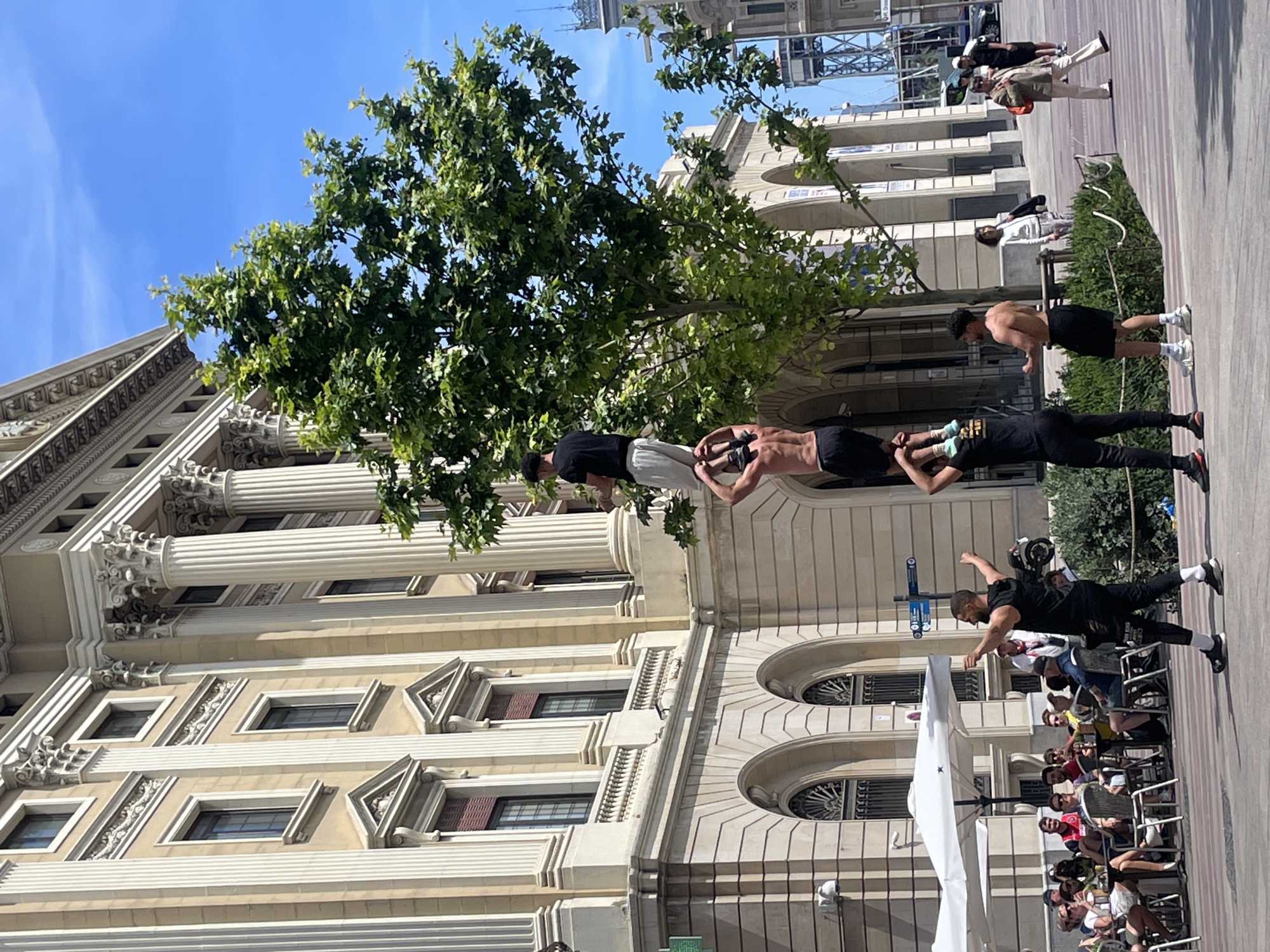 The image shows a street performance taking place in Barcelona, Spain. Three men are engaged in an acrobatic feat, with one man lying on the ground, another man doing a push-up on the feet of the man on the ground, and a third man standing on the back of the man doing a push-up. They are performing on a pedestrian walkway lined with chairs, where a crowd of people are seated, observing the performance. The scene is set against a backdrop of a large, ornate building with classical architectural elements, including columns, arches, and decorative moldings. A green tree is partially obscuring the building. The sky is blue and clear.