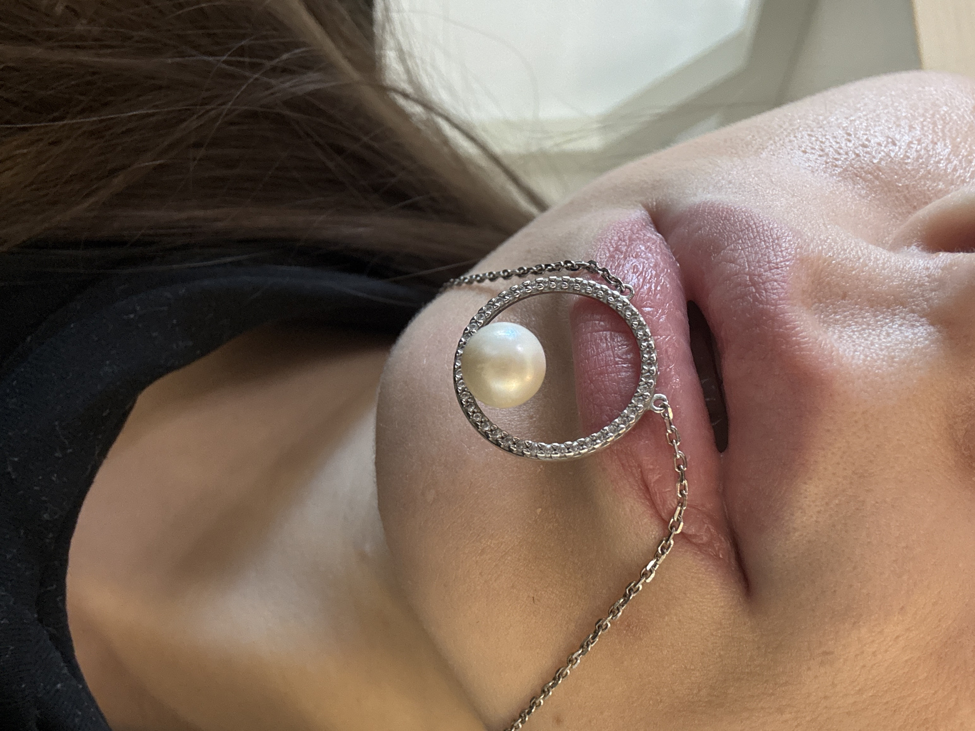 The image shows a close-up of a person's lips with a necklace adorned with a pearl and small diamonds. The necklace is positioned around the mouth, with the pendant hanging just below the lower lip. The person's skin is visible, with some texture and pores. The background is blurred, suggesting an indoor setting. The image does not contain any information about the location in the city of Unknown, Russia.