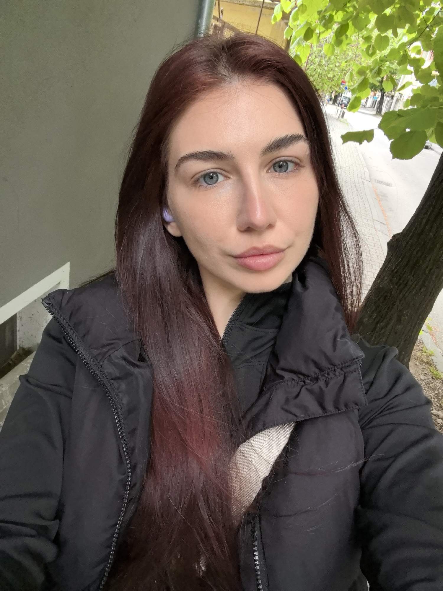 A woman with long, burgundy hair and fair skin is taking a selfie outdoors. She has light blue eyes and is wearing a black puffer jacket. In her left ear, she has a light purple earbud. The background includes a grey wall and a tree trunk with green leaves. Behind the tree, a street scene is visible, suggesting an urban location. This photo was taken in Stara Zagora, Bulgaria.
