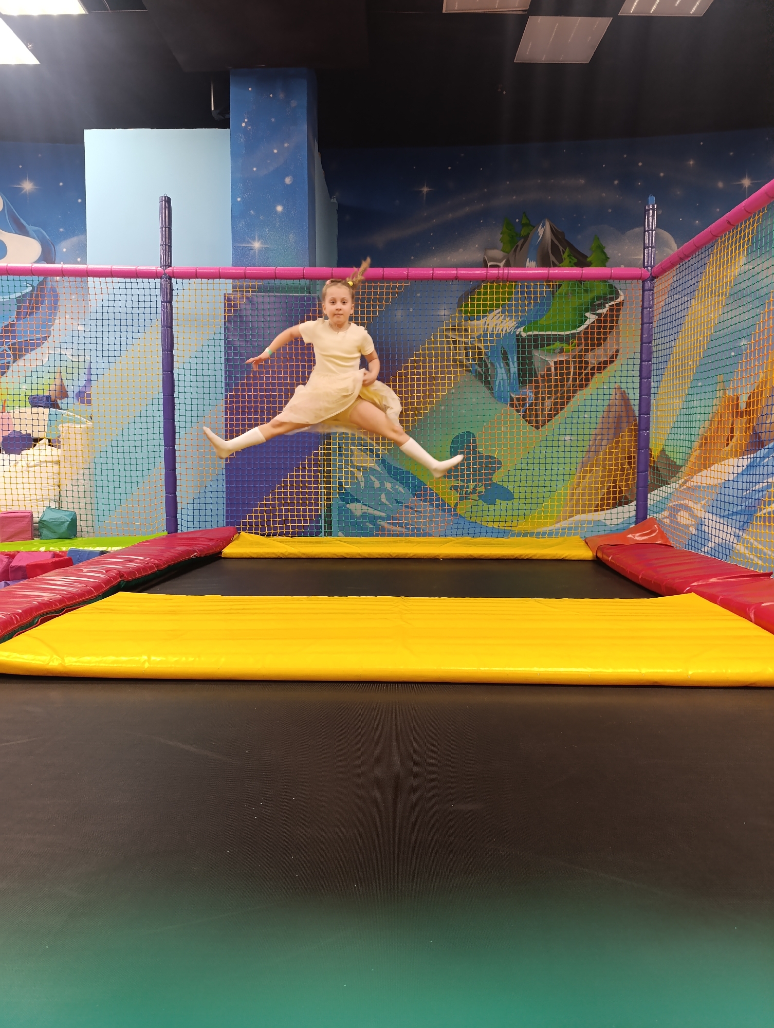 A young girl is jumping on a trampoline in an indoor playground. The trampoline has a black jumping surface, yellow safety padding around the edges, and a surrounding net with colorful supports. The girl is wearing a light-colored dress and white socks. The background features a wall with a mural depicting a landscape with mountains and a starry sky. The location is likely in Noginsk, Russia.