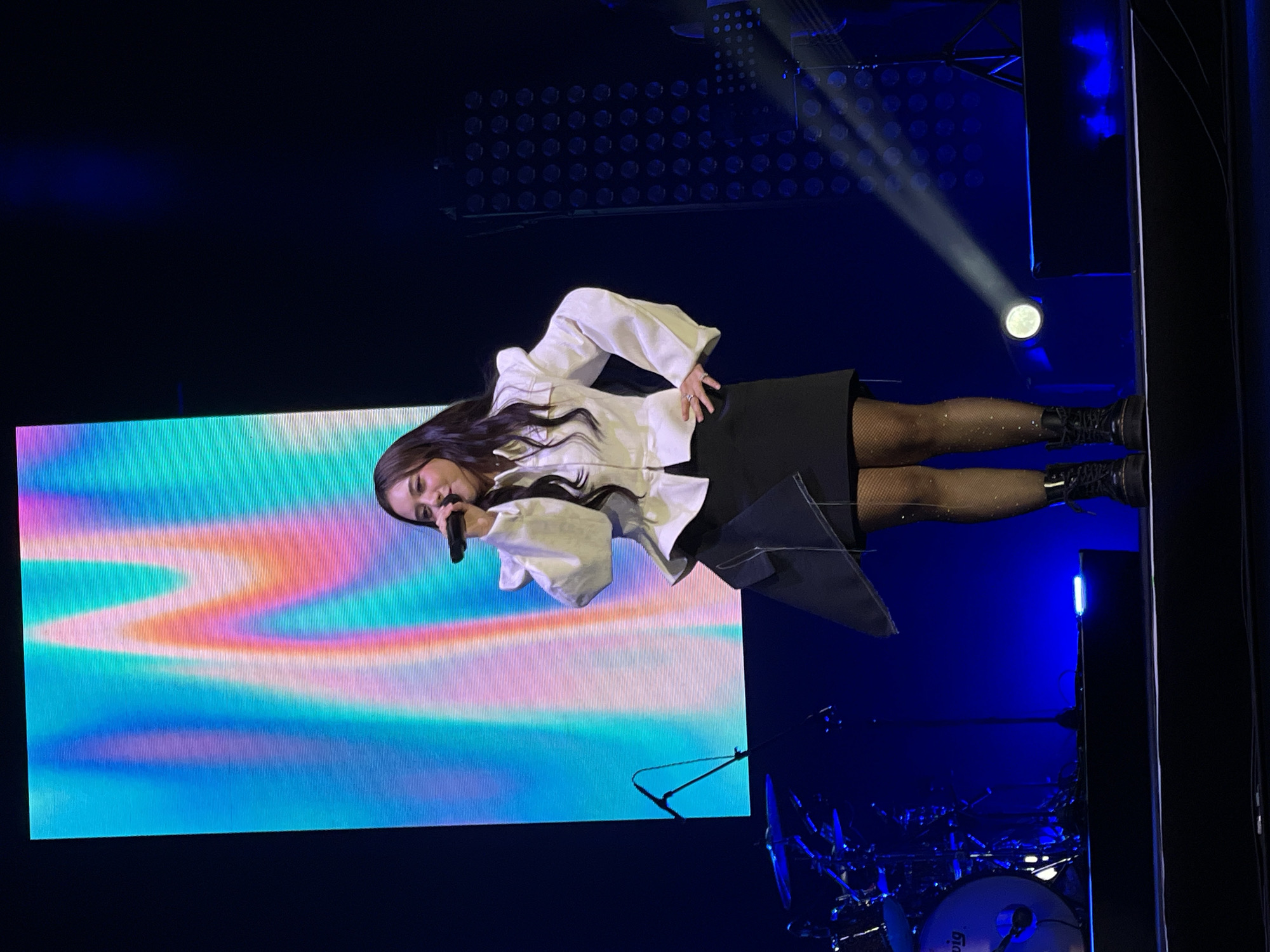 A woman is singing into a microphone on a stage. She is wearing a white blouse with long, ruffled sleeves and a black skirt. She is also wearing black tights and black boots. A large screen behind her displays a colorful, wavy pattern. Stage lighting is visible in the background. It is not possible to confirm if the location is Salembaran, Indonesia, from the image.