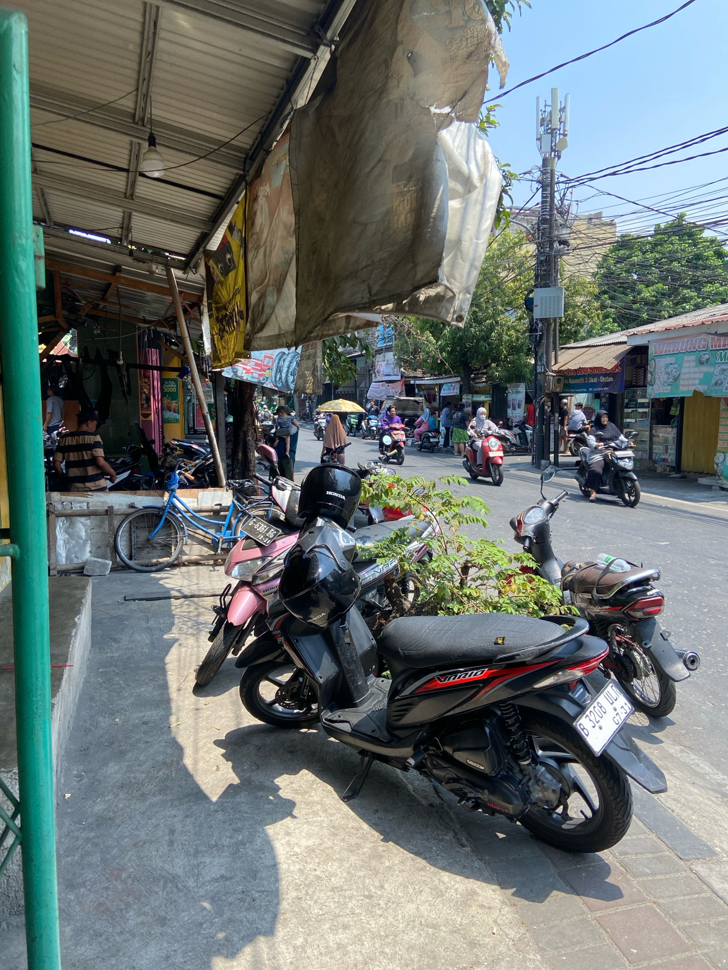 The image depicts a street scene in North Jakarta, Indonesia, on a sunny day. In the foreground, several motorbikes are parked on the sidewalk, with one black Honda Vario in sharp focus. A blue bicycle is also visible. The sidewalk is made of concrete and pavers, and it is bordered by a green pole on the left.
Behind the parked vehicles, a small shop or stall is visible, with a corrugated metal roof and various items displayed. A person in a striped shirt is standing inside the stall. Further down the street, traffic can be seen, including more motorbikes with riders, some of whom are wearing headscarves. A cell tower with multiple antennas stands prominently on the right side of the image, with numerous overhead wires. Buildings line the street, some with awnings and signs, suggesting a commercial or mixed-use area. The overall environment appears to be a bustling urban street with a mix of residential and commercial activity. The sky is clear and blue, indicating good weather conditions.