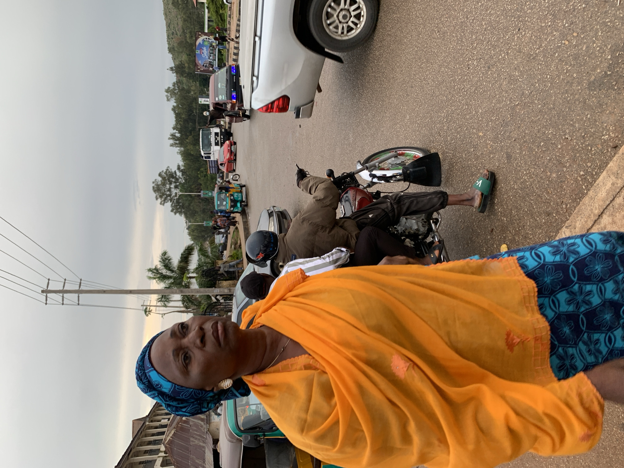 A medium shot captures an elderly African woman in the foreground, facing left and looking upwards with a pensive expression. She wears a vibrant orange draped scarf over a blue and black patterned skirt. A delicate gold chain is visible around her neck. Behind her, a man wearing a helmet and a brown jacket rides a motorcycle, and another person with a striped shirt is partially visible. In the background, an auto-rickshaw and a silver pickup truck are parked on a street with other vehicles and people. The scene appears to be a busy intersection in Gura Top, Nigeria, with buildings and trees lining the road under an overcast sky.