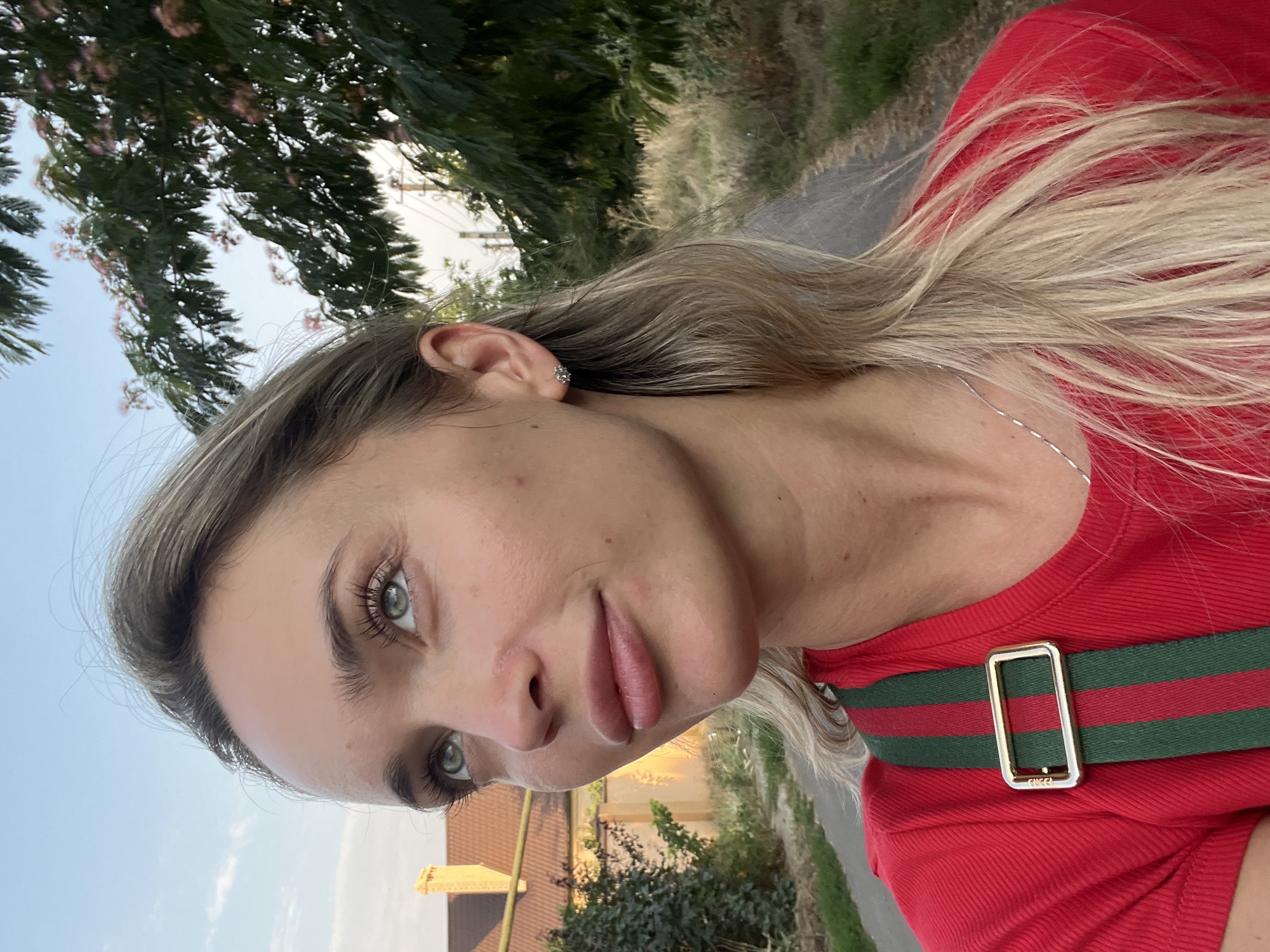 A woman with blonde hair is looking up and to her left. She is wearing a red shirt with a green and red striped strap with a gold buckle. She has a silver necklace. In the background, there is a house with an orange roof and a tree with pink flowers. The sky is blue with some clouds. The woman is positioned in the foreground, with the background blurred. The image appears to be taken outdoors in a residential area. The location is specified as Fontanka, Ukraine.