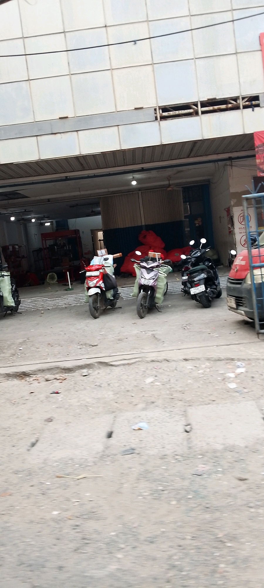 In Bekasi, Indonesia, a street scene captures multiple motorbikes parked in front of a building with a light-colored, tiled facade. Some motorbikes are covered with light-colored tarps, while others are uncovered. One motorbike in the foreground is red and white, another is dark colored with a purple tarp, and a third is black. Behind the motorbikes, inside a covered area, are stacks of large red objects, possibly fabric or bags. The ground in front of the building is paved and appears uneven, with some debris scattered about. The building itself has a corrugated metal overhang, and lights are visible inside. A red vehicle is partially visible on the right side of the frame, and a blue metal fence structure is also present. The overall impression is one of a busy commercial or storage area.