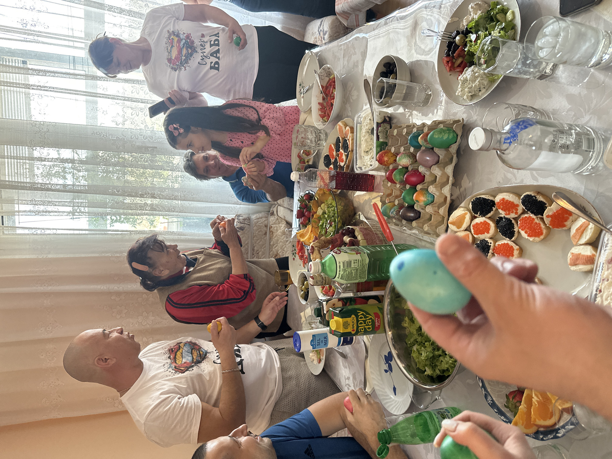 Here's a detailed description of the image:
**Location:** The scene appears to be set indoors, possibly in a home in Rakovski, Bulgaria. This is inferred from the Cyrillic text seen on a woman's T-shirt that reads "Супер БАБА" (Super Grandma), and the general ambiance.
**Scene:** The image captures a family gathering, seemingly during a festive occasion, possibly Easter, as evidenced by the presence of dyed eggs. A large table is the focal point, laden with a variety of dishes, including salads, sandwiches topped with caviar, fruits, and other snacks. There are also beverages, including bottles of juice and what appears to be a bottle of vodka.
**People:** Several individuals are present around the table. A man with a shaved head wears a white T-shirt featuring a Superman logo with what seems to be traditional clothing. A woman with short hair sits nearby, wearing a red shirt. A younger girl in a pink dress sits next to a man in a blue shirt, they are also participating in the activities. A woman in a white shirt featuring Cyrillic text stands near a window, possibly taking photos with her phone. Arms and hands reach towards the table, indicating people are interacting with the food and each other.
**Objects:** Besides the food and drinks, the table also features an egg carton filled with dyed Easter eggs. There are plates, glasses, and serving utensils scattered around the table. A window with translucent curtains can be seen in the background, providing natural light.
**Actions and Interactions:** The individuals are engaged in conversation and eating. Some are holding colored eggs, possibly participating in the traditional Easter egg cracking game. The overall atmosphere suggests a joyful and familial gathering.