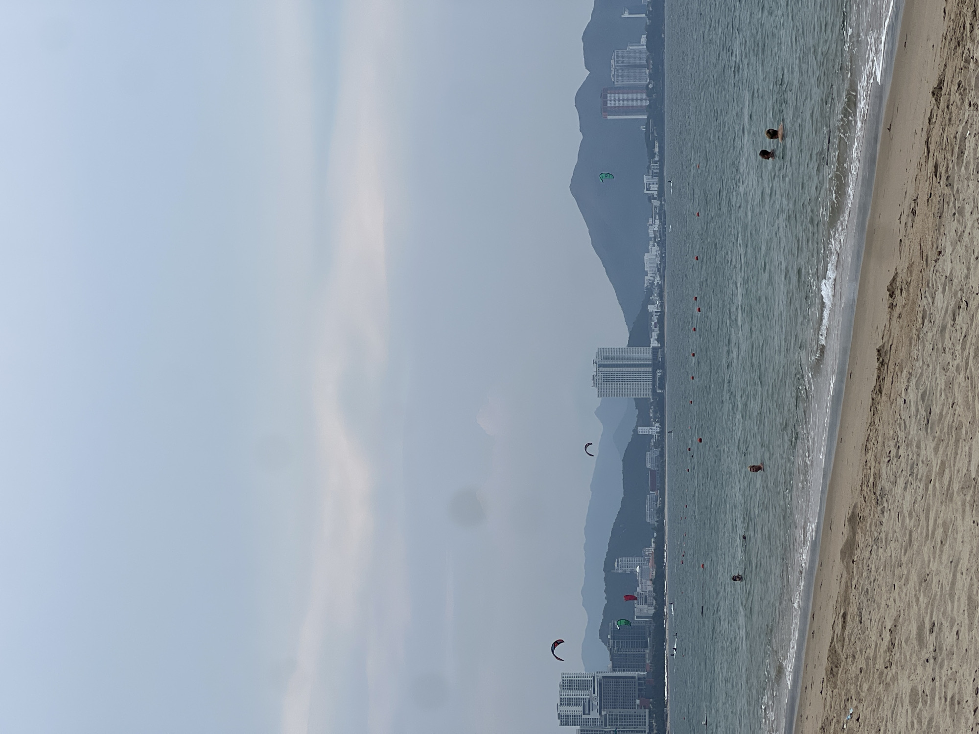 This panoramic image captures a coastal scene in Nha Trang, Vietnam, under an overcast sky. The shot is taken from a beach, looking across the ocean towards a city and mountains in the distance.
On the right side of the foreground, a wide stretch of light brown, sandy beach is visible, with gentle waves breaking at the water's edge. The mid-ground features the vast, calm, grey-blue ocean. Several people are swimming or wading in the water, with two individuals prominently in the central-right area. Red buoys are spaced out in the water, likely marking a swimming area or a boundary. High above the water, numerous colorful kites—including distinct green and red/orange ones—are airborne, indicating kitesurfing or kiteboarding activities.
The background is dominated by a bustling cityscape with a long line of modern high-rise buildings and skyscrapers extending along the coast. These buildings appear somewhat hazy, receding into the distance. Behind the urban sprawl, a range of large, dark mountains rises, their silhouettes softened by the atmospheric conditions. The sky is a uniform, pale grey, suggesting a cloudy or hazy day, which casts a soft, diffused light over the entire landscape.
The overall scene conveys a sense of leisure and recreation, with both relaxed beach activities and dynamic water sports taking place. No visible text is present in the image.