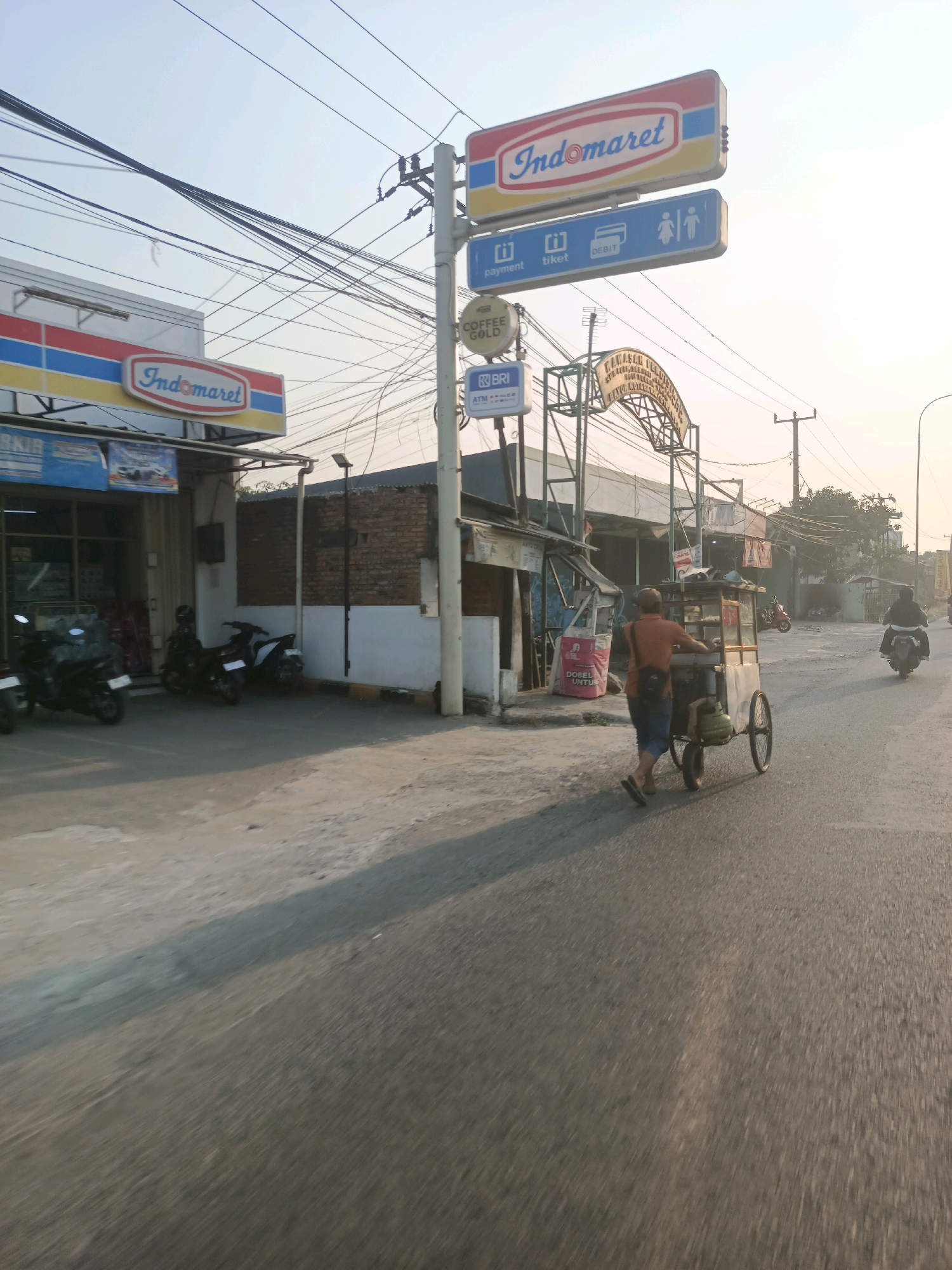 The image depicts a street scene in Karawang, Indonesia, likely in a commercial area. On the left, an Indomaret convenience store is visible with its distinctive red, yellow, and blue signage. Several motorcycles are parked in front of the store. A utility pole with numerous wires stands prominently in the center, supporting various signs, including an Indomaret logo, and directional signs indicating "payment," "tiket" (ticket), and "DEBIT," along with restroom symbols. Below these, signs for "BRI ATM" and "COFFEE GOLD" are also present. Further down the street, an arched gateway with text in Indonesian is visible, possibly marking an entrance or a specific establishment. In the foreground and middle ground, a man is pushing a street food cart with two wheels, laden with goods and a gas cylinder, along the asphalt road. Another person on a motorcycle is seen riding away in the background to the right. The sky is hazy and bright, suggesting it is either early morning or late afternoon. The overall environment appears to be a typical urban street with mixed commercial activity.