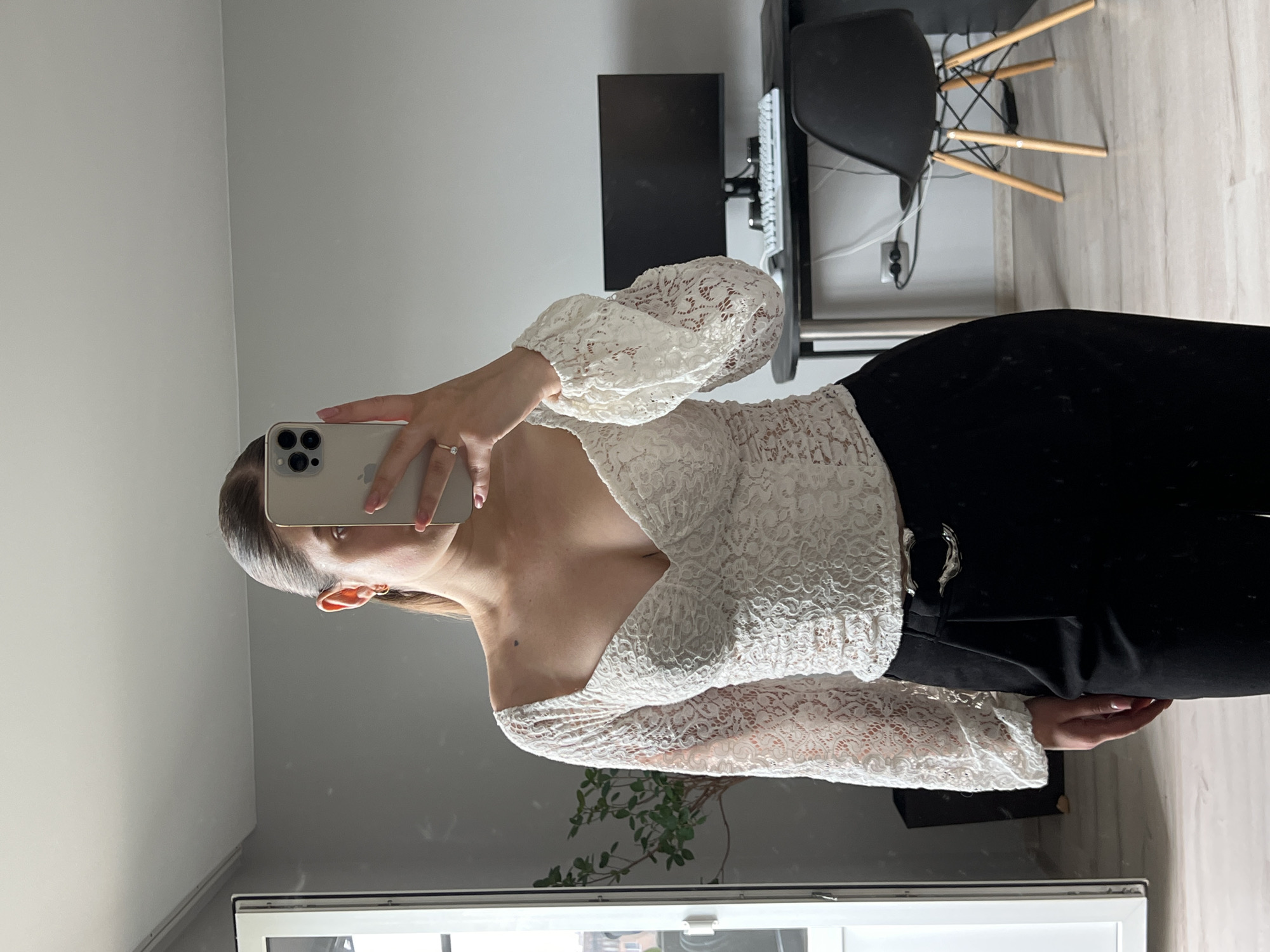 This mirror selfie captures a young woman posing indoors, likely within an apartment in Ternopil, Ukraine. The image is oriented vertically, rotated 90 degrees counter-clockwise from its natural upright position.
The subject is a female with light brown hair styled neatly, revealing a small hoop earring. She holds a light-colored smartphone, likely an iPhone given its triple camera array, to capture her reflection. Her nails are manicured with a light pink polish, and she wears a simple ring on her left ring finger. A small mole is visible on her chest.
She is dressed in a fashionable white top, characterized by an intricate lace or embroidered pattern and a flattering square or V-neckline. The top features voluminous, long sleeves that gather at the wrists. This is paired with black trousers or a skirt, secured at the waist with a distinctive silver-toned buckle.
The background, visible as a reflection in the mirror, depicts parts of a contemporary room. To the left is a plain light grey wall. In the upper right corner, a desk with a black computer monitor and a white keyboard is visible. A modern black chair with wooden legs stands behind the desk. Towards the bottom, a section of a white-framed window reflects an outdoor scene that appears to be urban. A small potted plant with green foliage is also reflected near the window. The floor is covered with light-colored wooden or laminate flooring. The overall lighting suggests it is daytime. No visible text is present in the image.