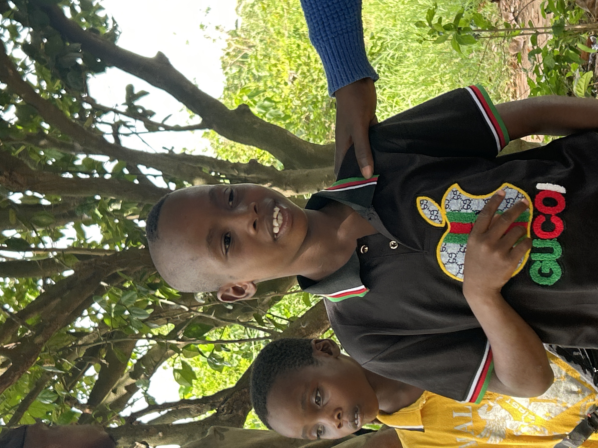 The image shows two young Black boys outside in a natural setting, likely a park or wooded area, with trees and foliage visible in the background. The taller boy, closer to the camera, is wearing a dark-colored polo shirt with a Gucci logo and red, white, and green striped trim. He is smiling and looking towards the camera. A hand with a blue sleeve is touching his shoulder. The second boy, in the lower portion of the frame, is wearing a yellow t-shirt. The location is specified as Unknown, Unknown.