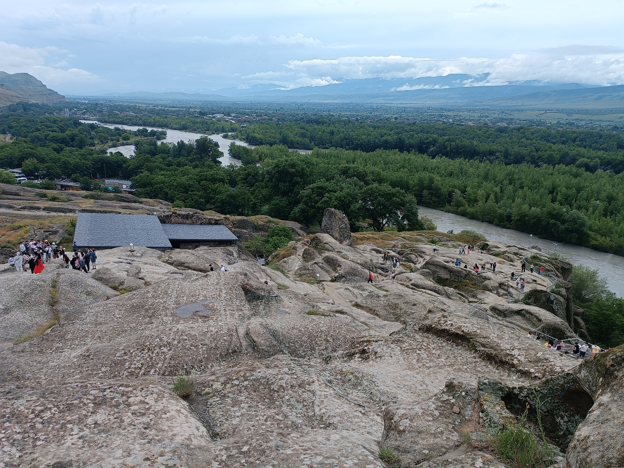This is a wide-angle, high-angle, eye-level shot of the ancient cave city of Uplistsikhe in Georgia. The foreground is dominated by rough, weathered, beige-colored rocks with patches of lichen. In the middle ground, a group of tourists is visible on the rocks, some walking and others standing. To the left of the tourists is a dark grey, gabled roof structure, possibly a visitor center or museum.
Beyond the rocky terrain, a wide, muddy brown river flows, bordered by a dense, dark green forest. The forest stretches into the distance, where a hazy landscape of hills and mountains can be seen under a cloudy, overcast sky. The overall impression is of a historical site integrated into a natural landscape.