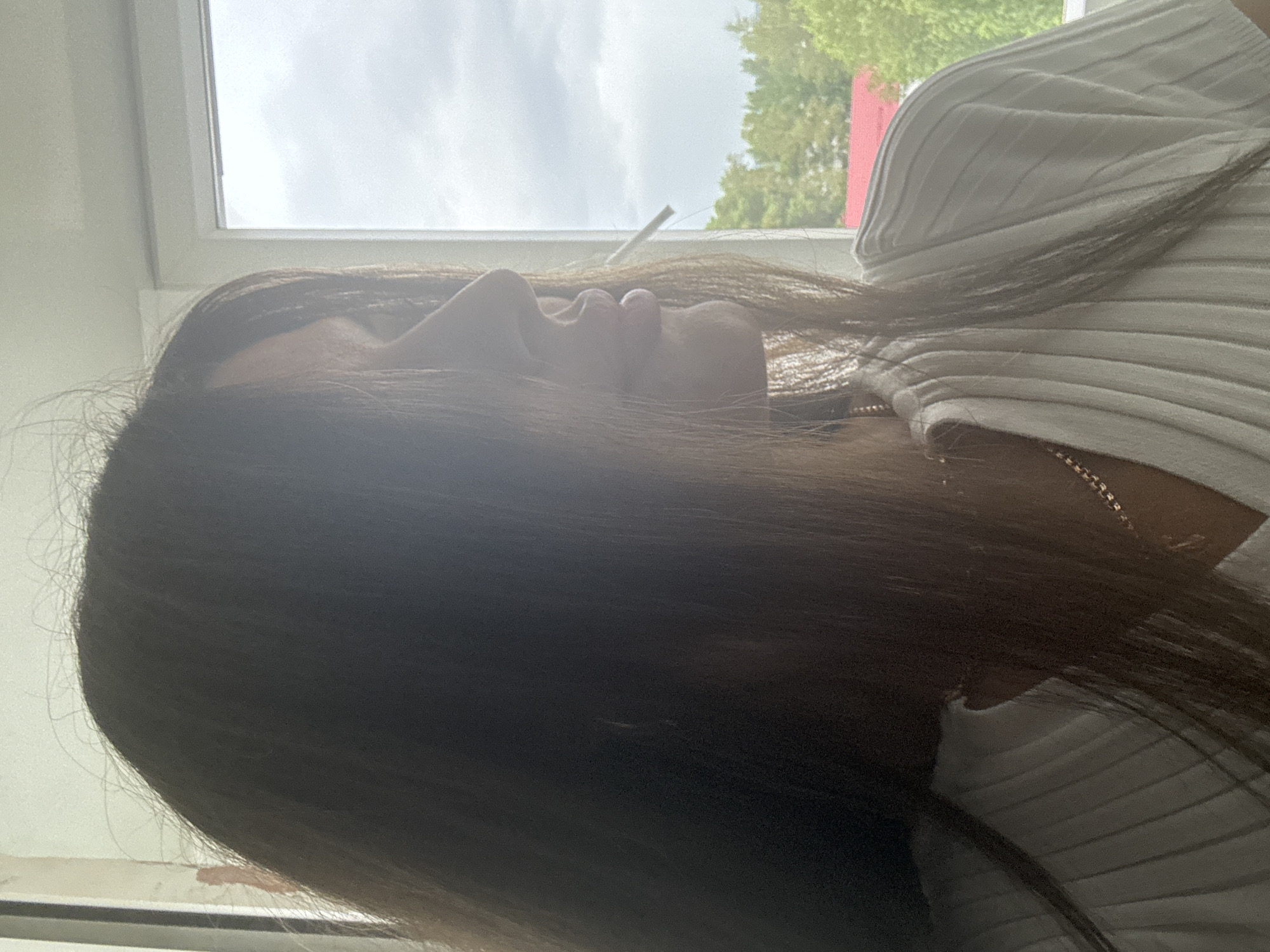 The image shows a close-up profile view of a young woman looking out a window. She has long, dark brown hair that partially obscures her face, and her head is tilted slightly upwards. She is wearing a white ribbed shirt and a gold chain necklace. Her right hand appears to be holding a cigarette, which is visible between her lips. The background shows a window with a cloudy sky and greenery outside, including trees and a red-roofed building. The lighting suggests it is daytime, possibly overcast. The image was taken in the city of Chaikovsky, Russia, as indicated by the metadata.