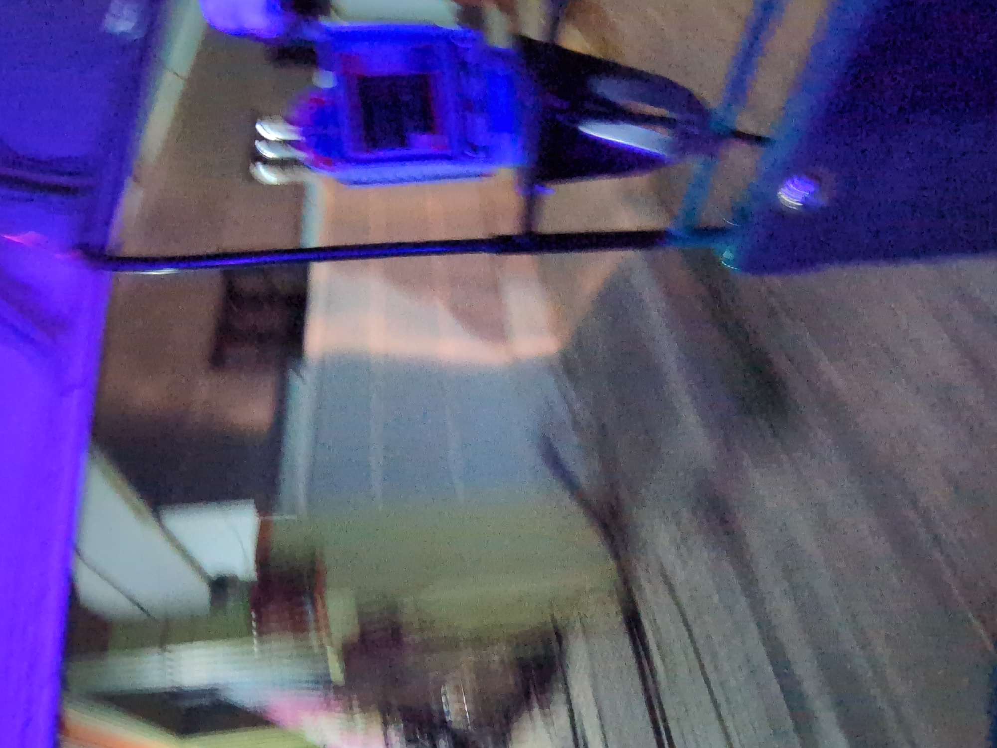 The image is a severely blurry, noisy, and abstract photograph dominated by intense artificial blue and purple lighting. The primary subject, if the image is mentally rotated 90 degrees counter-clockwise, appears to be a technological device, possibly a drone or a component of one, positioned in the upper central area. This object is primarily blue/purple, suggesting either its material or, more likely, illumination by strong blue lights, possibly LEDs. Within this main object, several small, bright metallic or white elements are visible, which could be connectors, sensors, or reflective parts. A thin, dark bar extends horizontally across the frame, seemingly connected to the main blue object. To the right, a larger, indistinct mass of dark blue or purple is present, which might be another piece of equipment or part of the same setup.
The background is extremely out of focus, making it challenging to identify specific details. It suggests an indoor or semi-enclosed setting with very low ambient light, contrasting with the bright blue illumination of the subject. Vague, blurred shapes that could be walls, floors, or other structures are discernible.
There are no visible people, discernible activities, or legible text. The image's quality and heavy reliance on artificial lighting suggest a focus on the aesthetic presentation or demonstration of the technological object. Given the context of Bengaluru, India, this could potentially be from a tech exhibition, product launch, or a laboratory setting, highlighting a piece of equipment under specific lighting conditions. The time of day is not evident, but the scene is artificially lit.