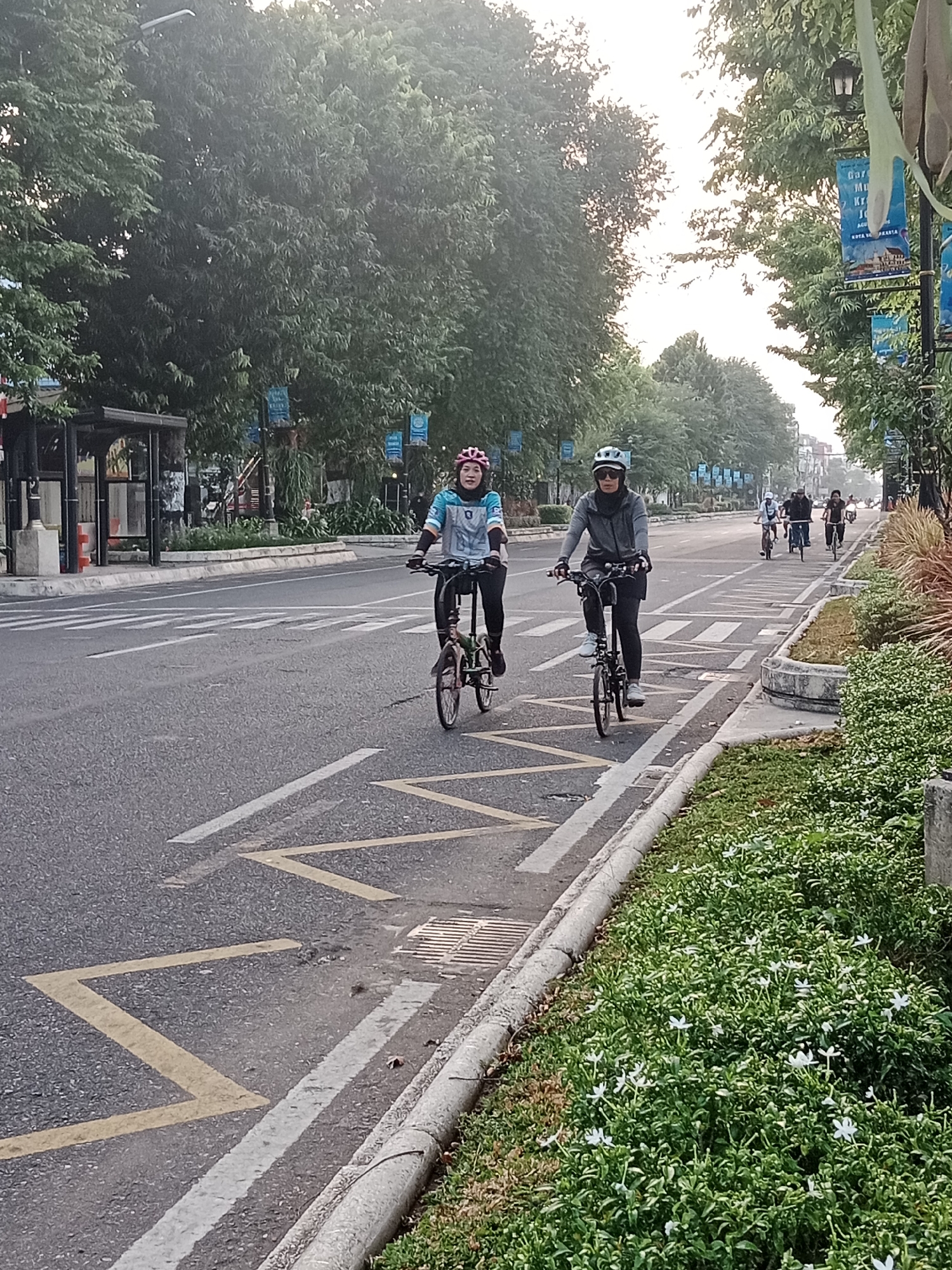 Two women are cycling on a paved road in Yogyakarta, Indonesia. They are both wearing helmets and athletic clothing. The woman on the left is wearing a pink helmet and a blue and white jacket, while the woman on the right is wearing a white helmet and a gray jacket. They are cycling in a designated bike lane. In the background, there are trees, banners hanging from lampposts, and a bus stop. The road is marked with yellow and white lines. The scene appears to be captured during daylight hours, possibly early morning or late afternoon, given the soft lighting and the presence of cyclists.
