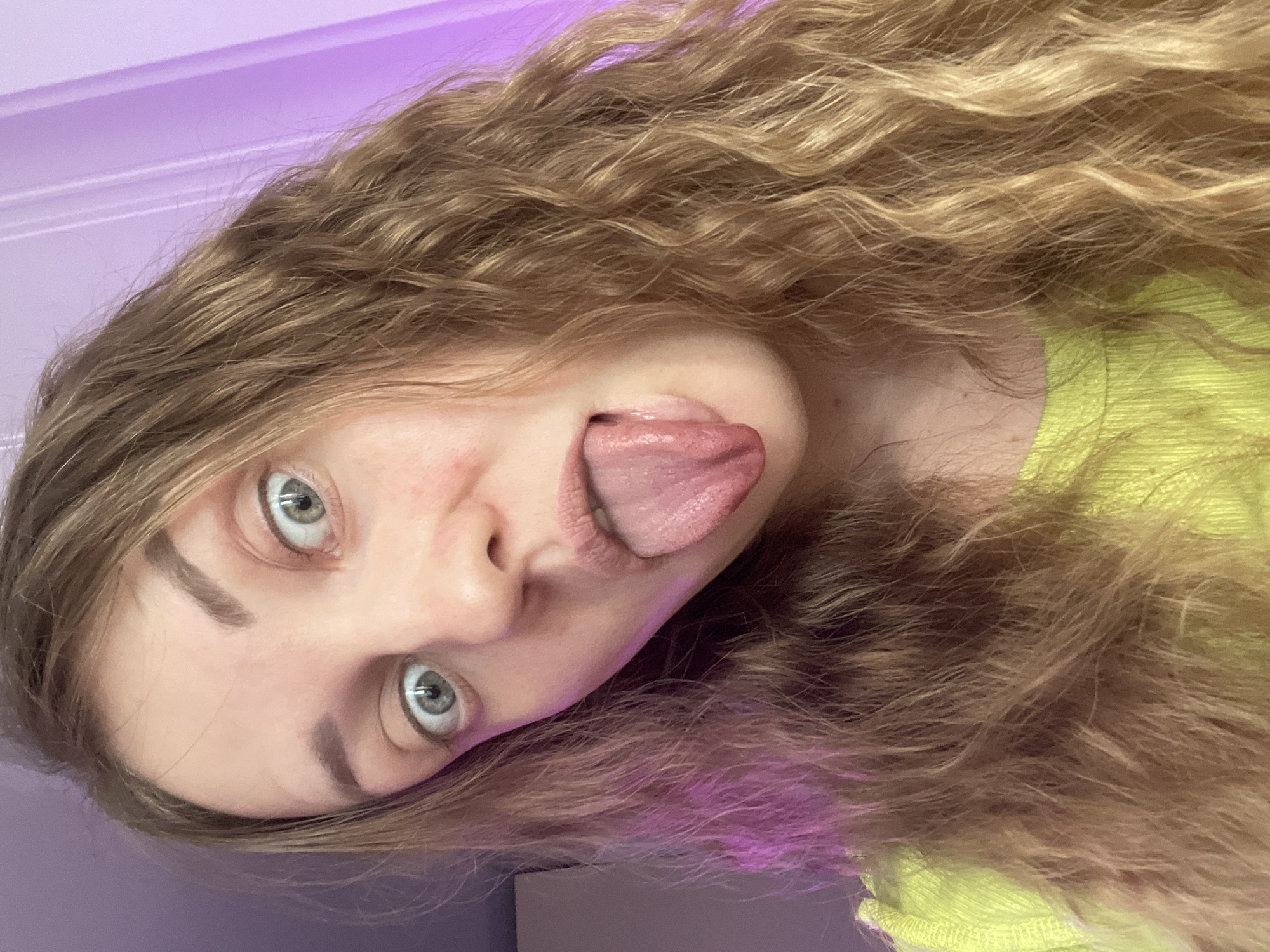 The image displays a close-up portrait of a single young female individual, showing her head and shoulders. She has long, wavy, light brown hair. Her blue-grey eyes are wide open, and her mouth is open with her tongue extended. She is wearing a lime green, ribbed garment. The background is an indistinct indoor setting, characterized by areas of purple and pink ambient lighting and a partial vertical white element, possibly a door frame or wall molding. No specific features are discernible within the frame to identify the location as being in the city of Batumi, Georgia.