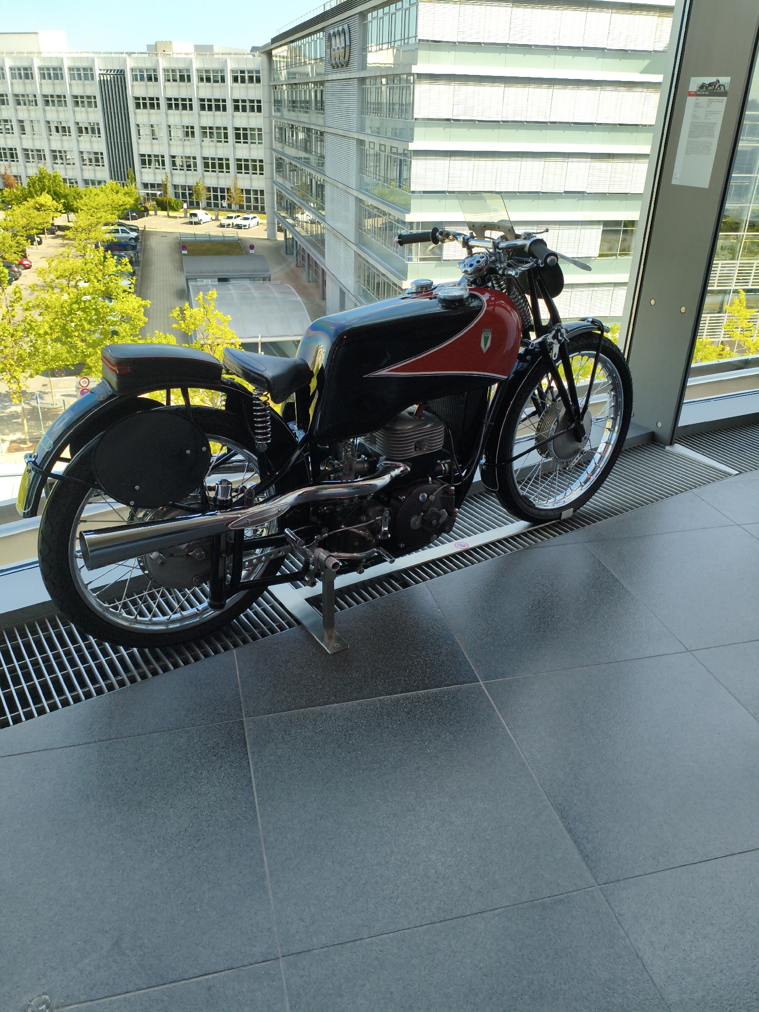 A vintage black and red motorcycle is displayed indoors, positioned on a metal grate next to a large window. Through the window, a modern office building with numerous windows is visible, along with trees and parked cars. The floor is tiled with large, dark gray square tiles. The scene is likely set in Ingolstadt, Germany, possibly at the Audi Forum Ingolstadt, which houses a museum showcasing Audi's automotive history. There are no people or actions depicted in the image.