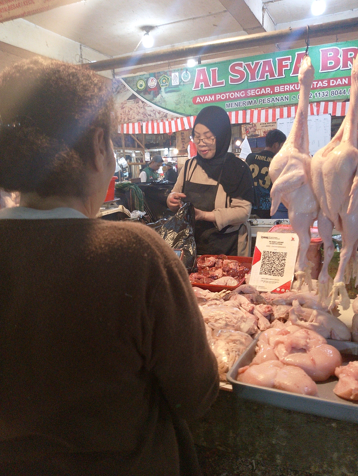 A mid-shot captures a butcher stall in Lembang, Indonesia, focusing on a woman in a headscarf and glasses. She wears a dark apron over a light-colored garment and is seen from the shoulders up, holding a black plastic bag. In front of her, a display of raw chicken parts and whole dressed chickens hangs from hooks. A red tray holds other raw chicken cuts. A QR code is prominently displayed on a white card. Behind the woman, a banner with red lettering reads "AL SYAFA BR..." and other text in Indonesian. Several other individuals are partially visible in the background, including a man wearing a numbered shirt. The environment appears to be an indoor market with a fluorescent light fixture illuminating the scene.