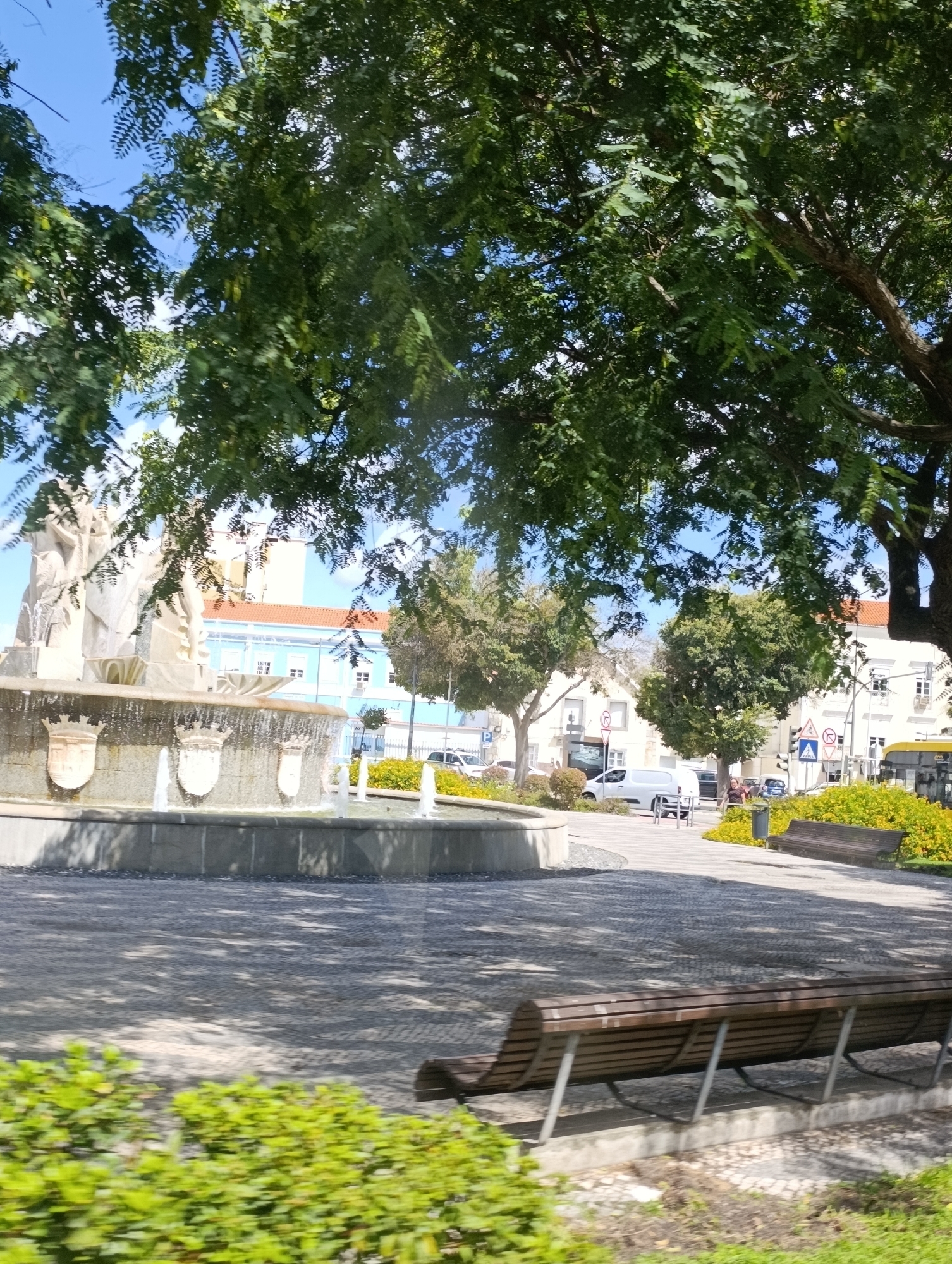 The image depicts a public square in Setúbal, Portugal, featuring a central fountain with multiple tiers and water spouts. The fountain is adorned with three crests or shields. Surrounding the fountain is a paved area with stone patterns, interspersed with patches of green foliage and benches. In the background, buildings with light blue and white facades are visible, along with a building with an orange roof. Trees with dense green canopies cast shadows on the ground. Several vehicles, including a white van, a car, and a yellow bus, are present in the square. A few individuals are seen walking or standing. The sky is clear and blue.