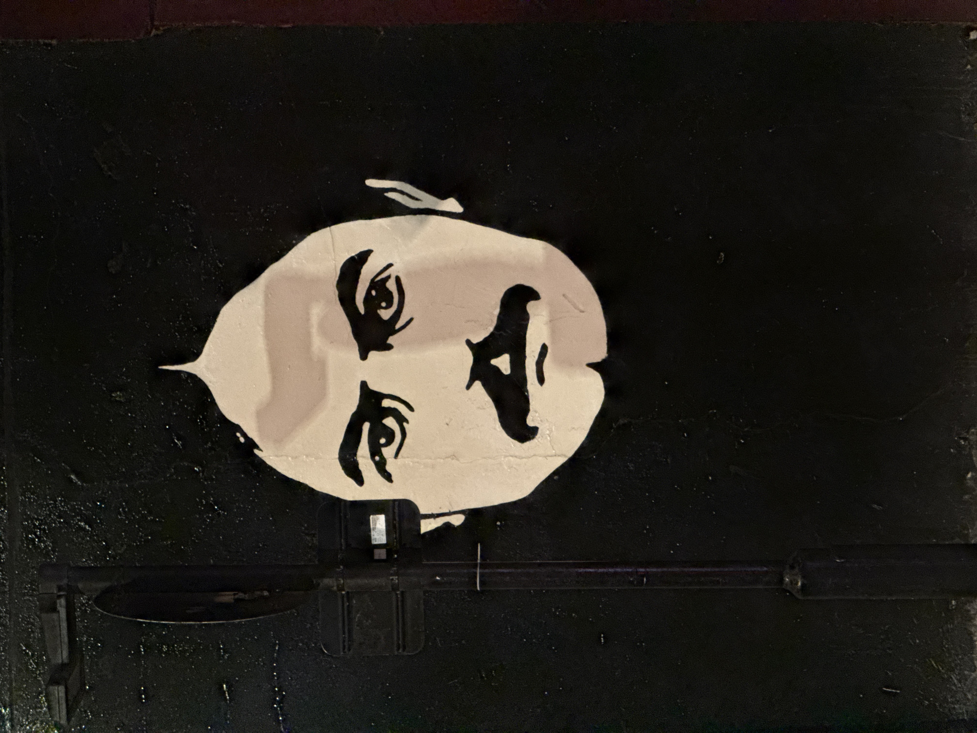 A black and white stencil-style mural depicting the face of the character Astro Boy, or Atom Boy, adorns a black surface. The head is depicted in profile. A metal latch mechanism is visible in the lower portion of the image. The piece can be found somewhere in the city of London, United Kingdom.