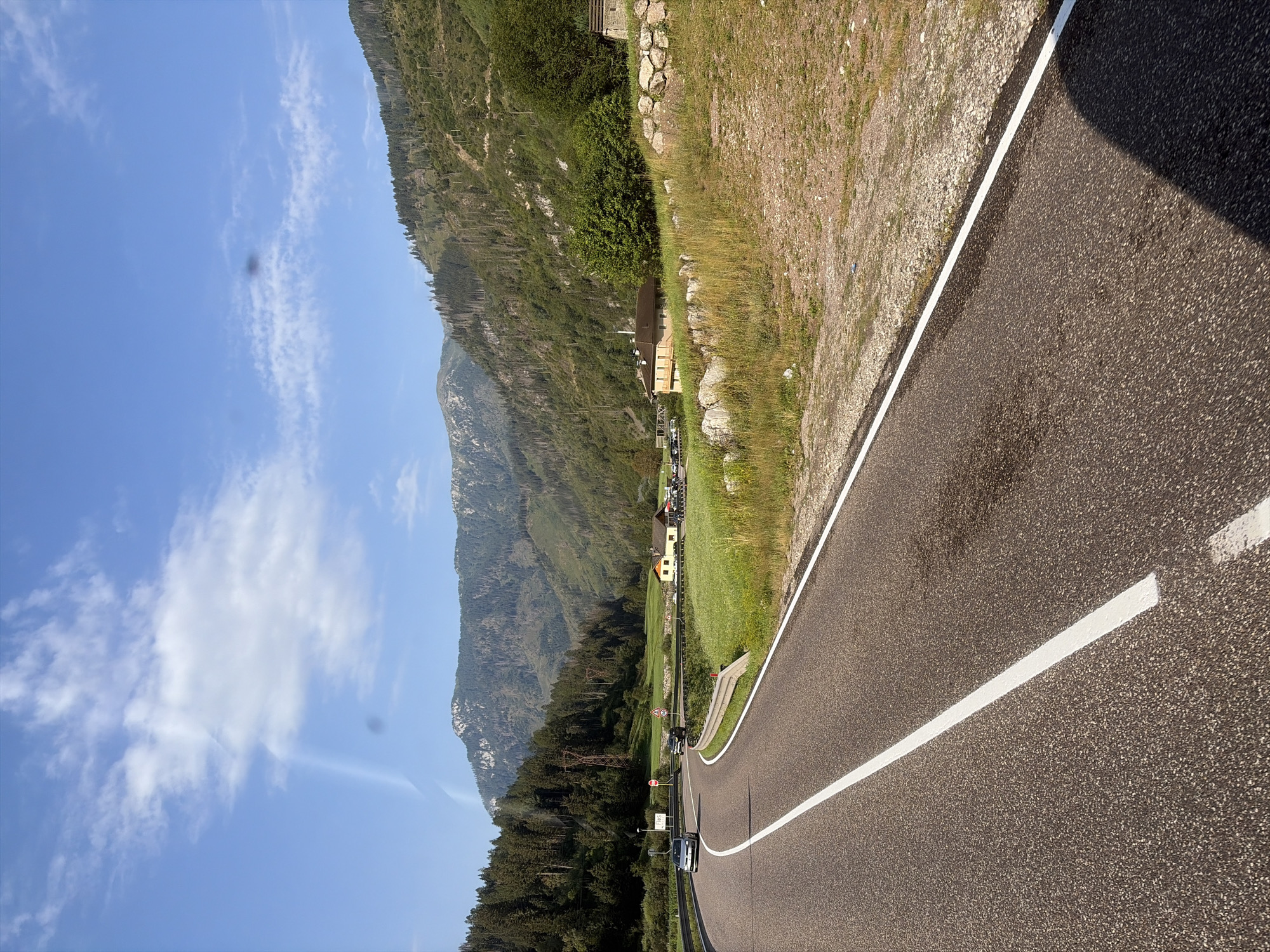 The image depicts a scenic mountain road in the vicinity of Moena, Italy, on a clear, sunny day. The asphalt road curves to the left, marked with a solid white line on the right edge and a broken white line on the left. Two cars are visible on the road: a dark colored sedan on the left and a black SUV further ahead.
To the right of the road, a grassy field slopes upwards, bordered by a stone wall. Beyond the field, a small cluster of buildings is visible, including a yellow house with a red roof and another tan building. In the background, a dense forest covers the lower slopes of majestic mountains, their peaks etched against a bright blue sky with scattered white clouds. The overall impression is one of a peaceful, rural landscape.