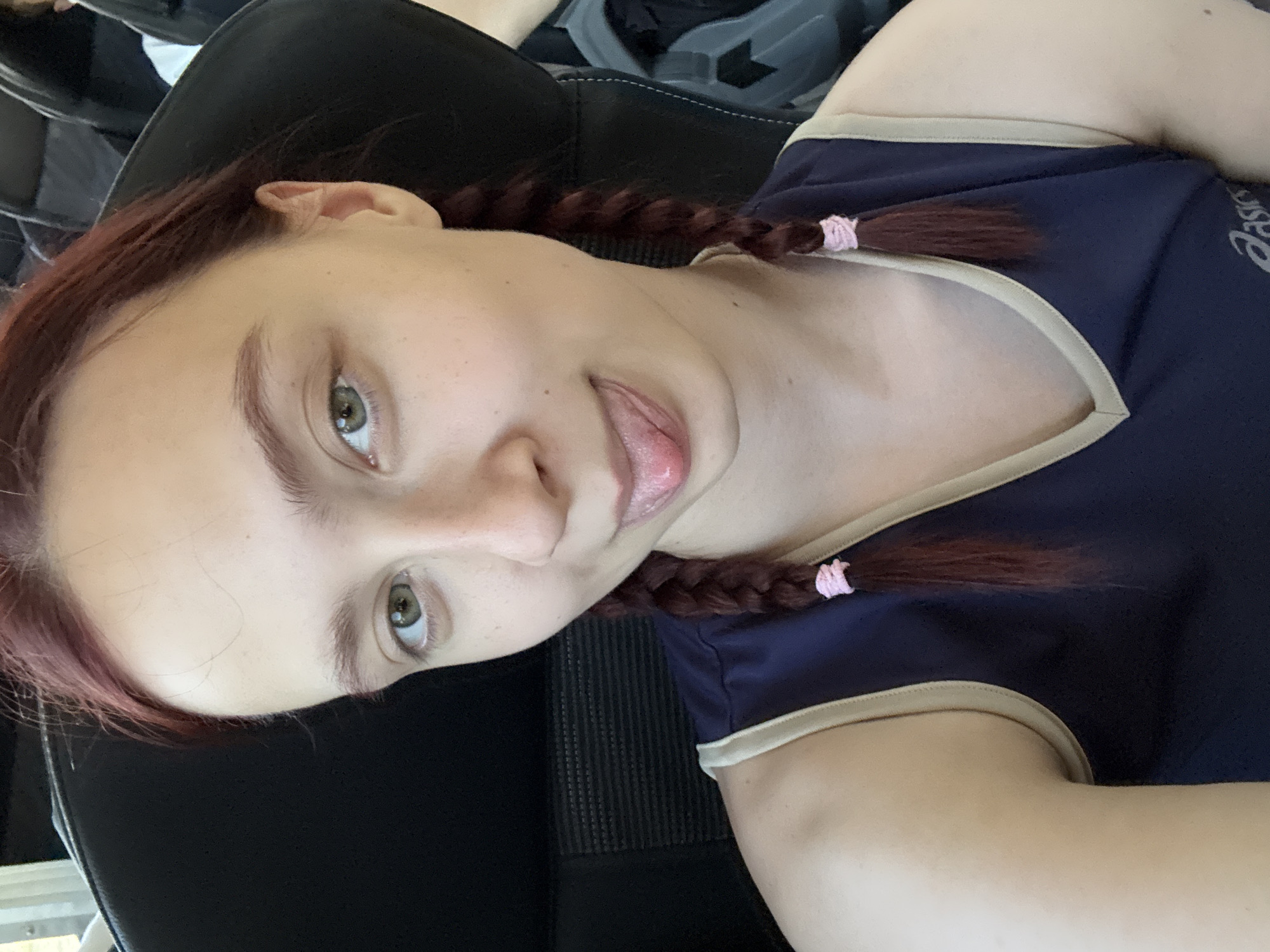 A young woman is seated in a black chair. She has light skin, green eyes, and reddish-brown hair styled in two braids secured with pink elastics. Her tongue is sticking out slightly. She is wearing a dark blue top with a tan trim and a small "Asics" logo on the right side of her chest. The background is blurry and unclear. This image does not include information about location in Votkinsk, Russia, or details about the environment.