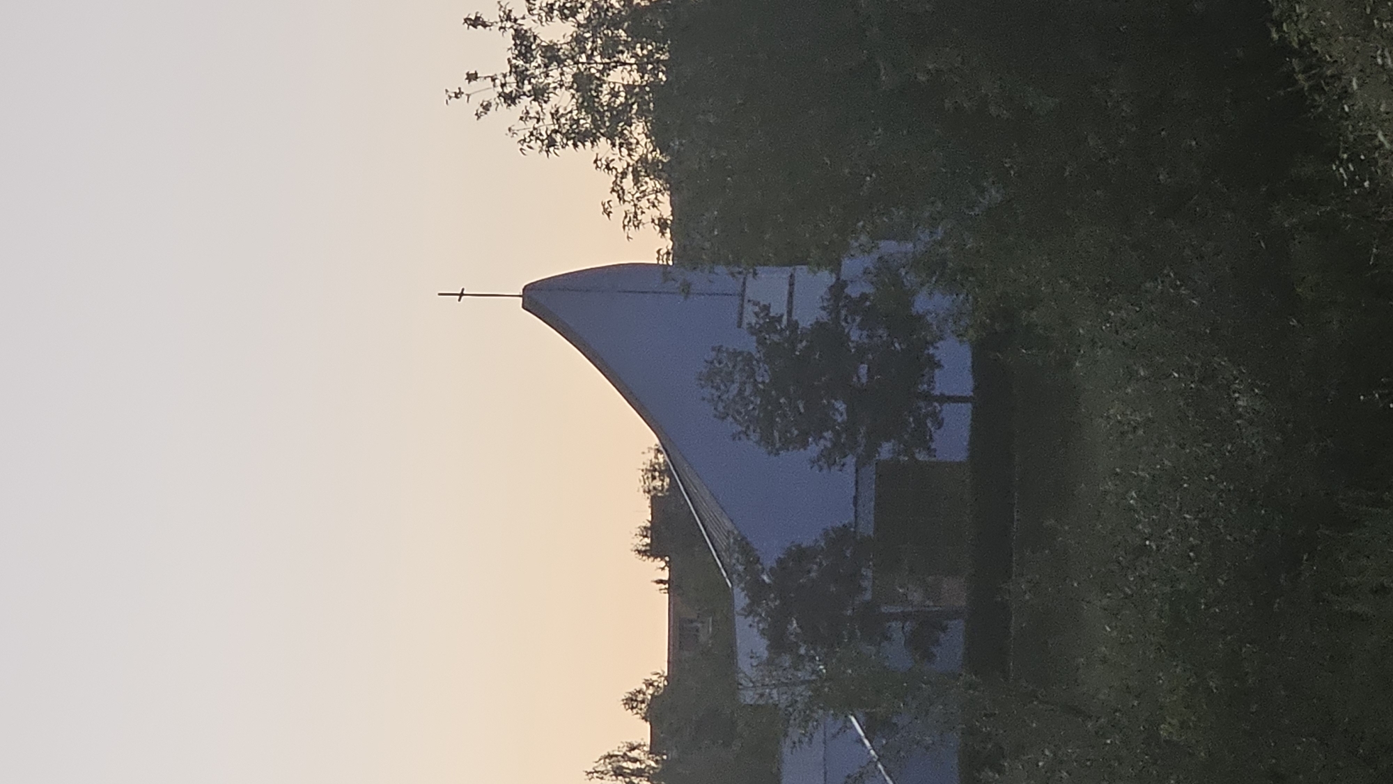 The image shows a modern, light blue church with a distinctive, sharply angled roofline, topped with a cross. The church is situated amidst lush green trees and foliage, with a hint of an urban landscape with buildings visible in the background. The sky is a soft gradient of pale orange and white, suggesting either sunrise or sunset. The overall impression is one of a serene and somewhat abstract architectural form against a natural backdrop. While the image does not provide specific geographic markers for Prešov, Slovakia, it captures a contemporary religious structure surrounded by nature.