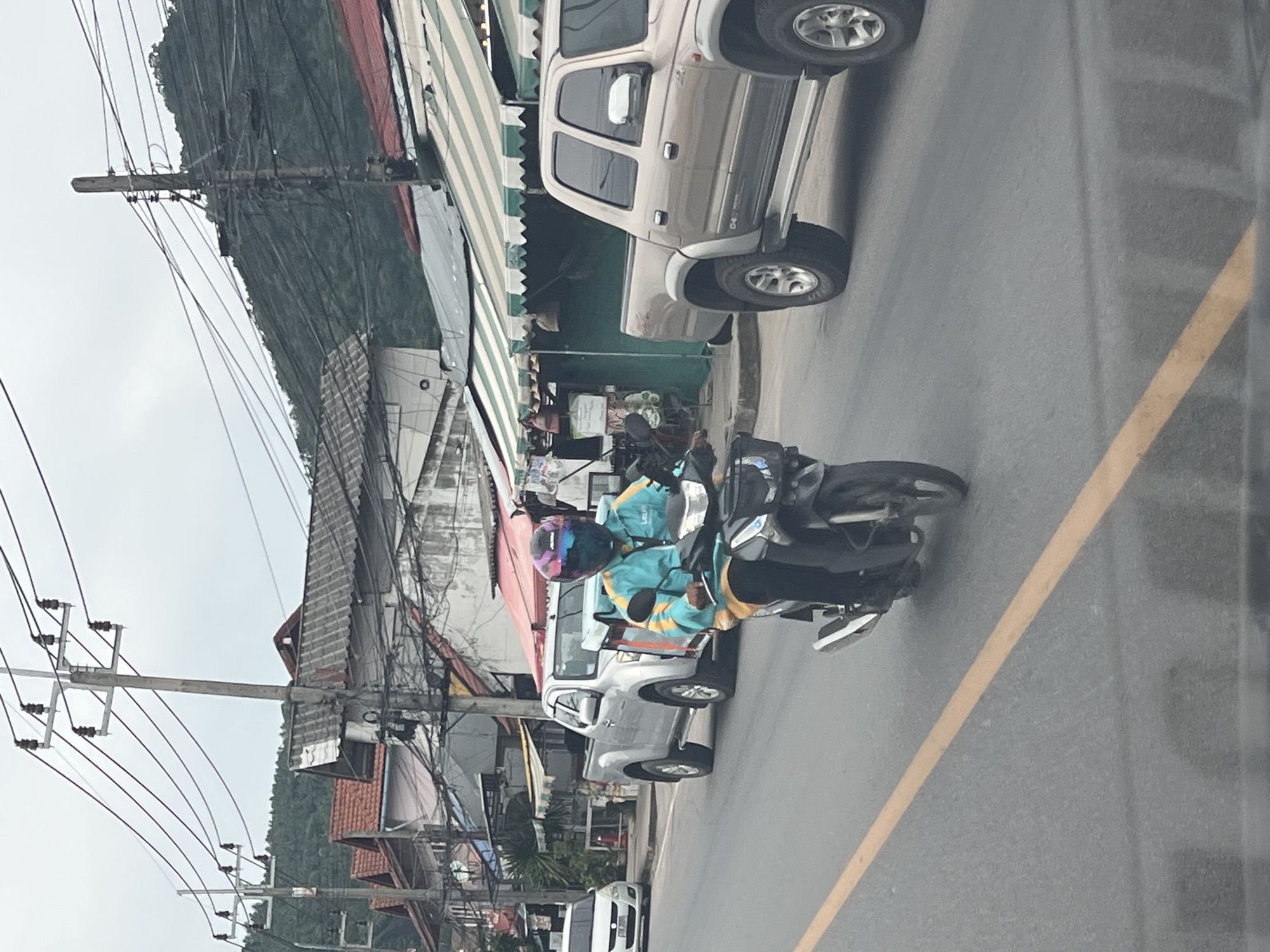 In Baan Hua Thanon, Thailand, a person rides a black motorcycle on the right side of a paved road, facing forward. The rider wears a bright blue and yellow jacket, dark pants, and a multi-colored helmet with a reflective visor. The motorcycle has a black body and chrome accents. Behind the motorcycle, a silver pickup truck is parked on the side of the road, and further back, a white car is visible. The background consists of buildings with tiled roofs, a green hillside with lush vegetation, and a cloudy sky. Electrical poles and wires are prominent features of the environment.