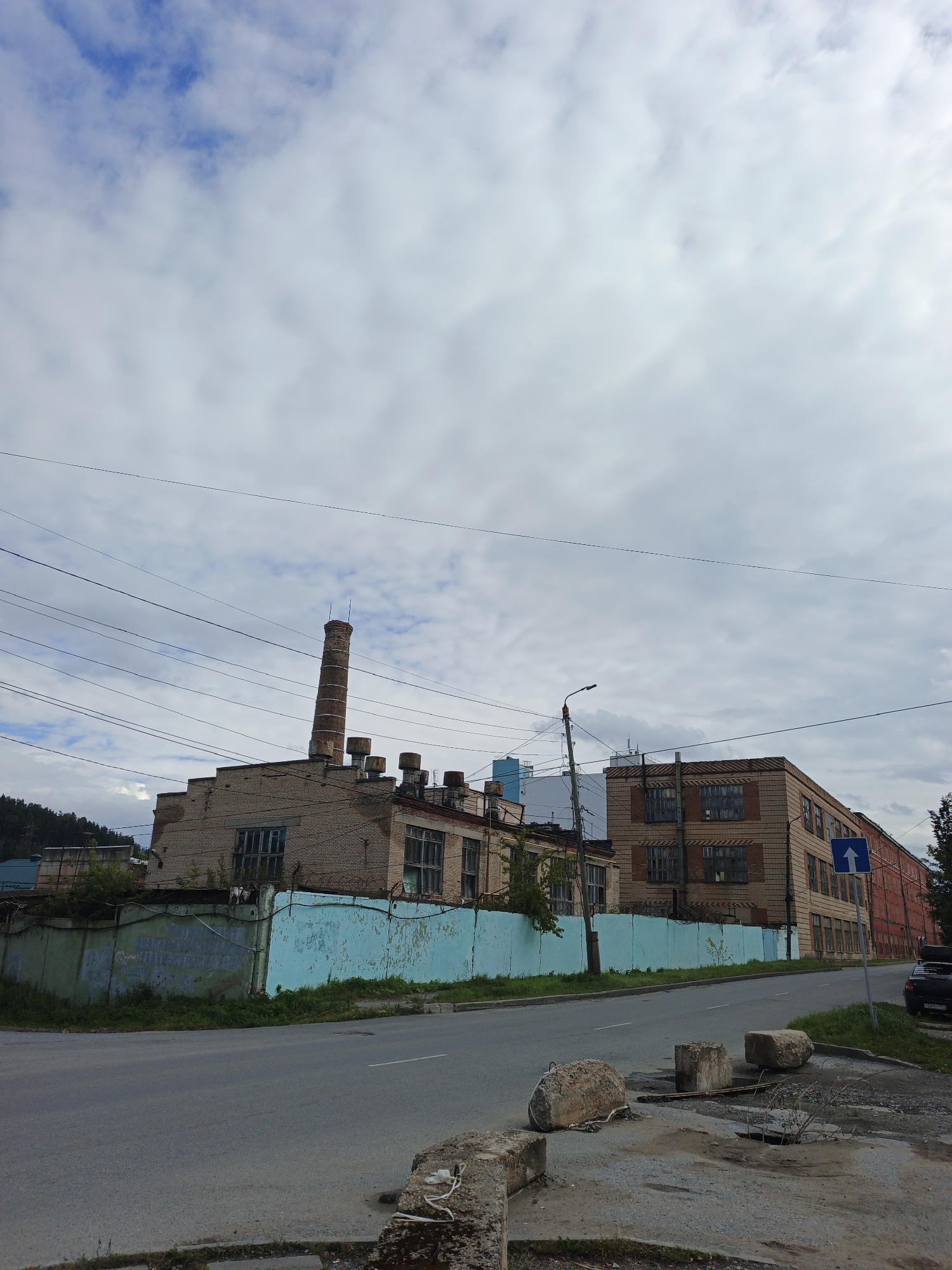 The image depicts an industrial area in the city of Zlatoust, Russia. In the foreground, a paved road curves to the left, with several large concrete blocks and some debris on the roadside. A blue and green fence runs along the edge of the road, bordering a complex of industrial buildings. The buildings are constructed of brick and have multiple windows. A tall, brick smokestack stands prominently next to one of the buildings. Overhead, a network of power lines crisscrosses the sky. The sky itself is partly cloudy with a mix of blue sky and white clouds. In the background, a wooded hillside is visible. A street sign indicating a one-way direction is posted on the right side of the road, near one of the buildings. A dark-colored car is parked on the right shoulder of the road. The scene appears to be a typical view of an industrial district, possibly showing signs of aging or disuse given the condition of the fence and roadside debris. There are no people visible in the image.