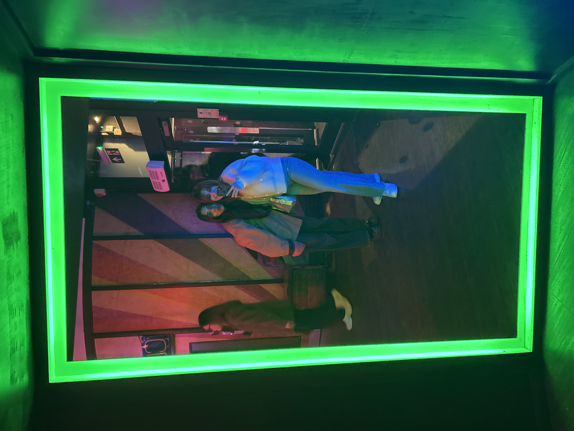The image shows a photograph of an illuminated green rectangular frame hanging on a dark wall. The frame contains a distorted reflection of the interior of a building. In the reflection, there are two people who appear to be posing for a photograph. One person is wearing a white jacket and jeans and the other person is wearing a green jacket and dark pants. There is also a reflection of another person in the bottom left corner. There are colorful diagonal stripes on the wall in the reflection and various objects like a container and signs can be seen in the background. The location is possibly in the city of London, United Kingdom, but this cannot be confirmed with certainty solely based on the image.