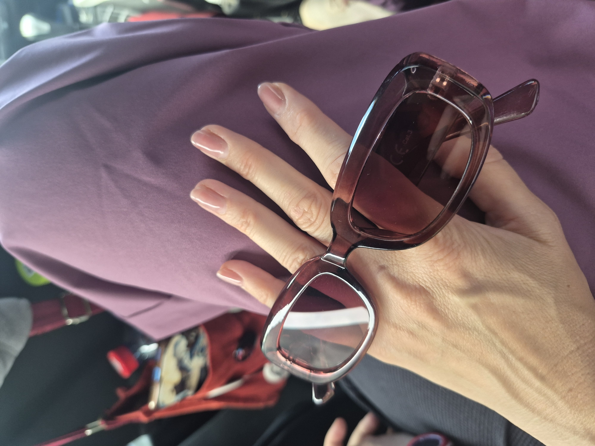 A person's hand, with neatly manicured nails, holds a pair of translucent rose-tinted sunglasses. The hand is positioned over a piece of mauve fabric, possibly clothing. In the background, partially visible, is a red bag with a strap, and a bottle of water. The scene appears to be indoors, possibly in a vehicle, given the blurred background elements which suggest movement. There is no specific information to indicate this image is located in Khrustal'nyy, Russia.