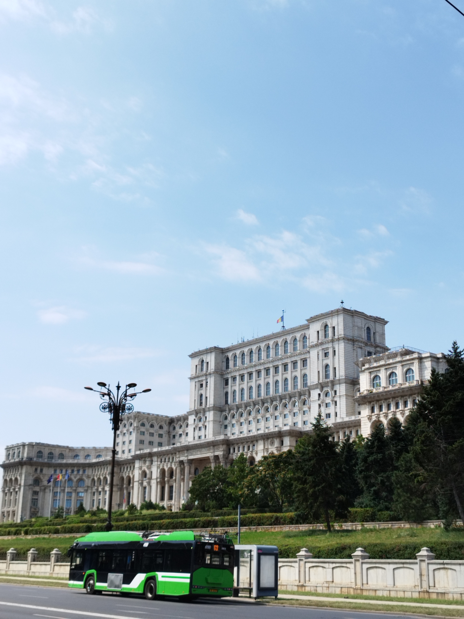 This is a full-frame, eye-level, outdoor, daytime, medium-shot photograph of the Palace of the Parliament in Bucharest, Romania, with a green and black public transport bus in the foreground. The sky is light blue with scattered white clouds. The Palace of the Parliament is a massive, imposing Brutalist building with a light-colored facade, characterized by numerous windows and arched entrances. A Romanian flag is visible flying from one of the upper levels. In front of the palace, there is a lush green landscape with manicured bushes and trees. A black street lamp with multiple lights is positioned between the bus and the palace. The bus is a modern, articulated electric bus, predominantly green with black windows and accents. It is stopped at what appears to be a bus stop, with a small, silver and grey bus shelter visible to the right of the bus. The bus has a route number "63" displayed on its front. The scene is captured from a street-level perspective, with a paved road in the immediate foreground.