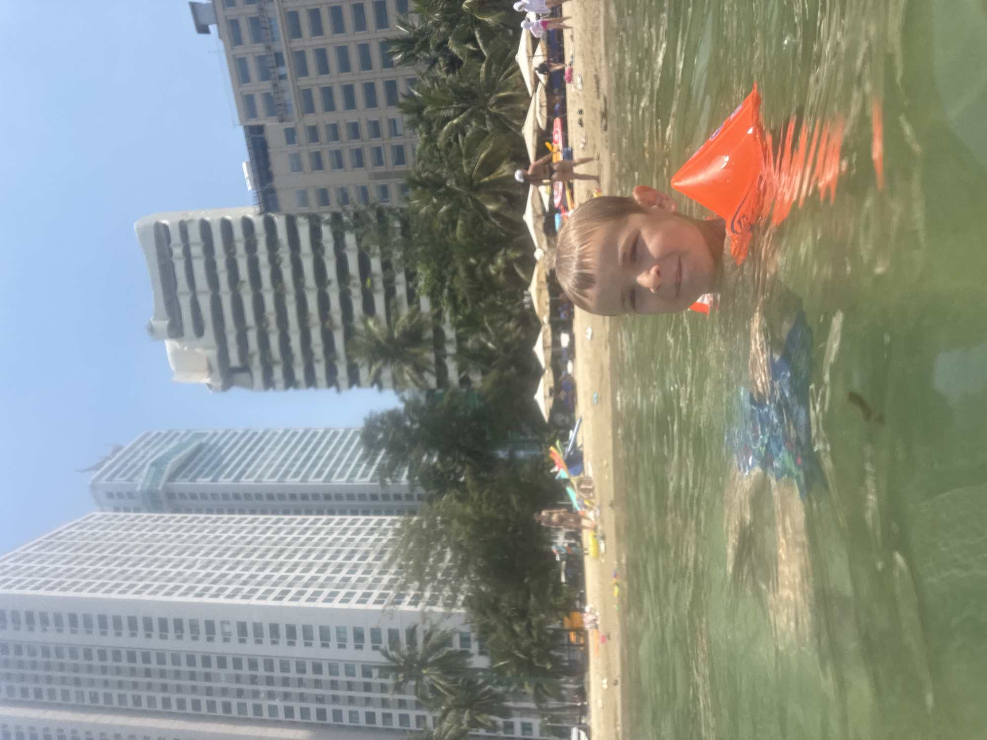 In the foreground, a young boy with short brown hair is partially submerged in the water, wearing blue swim trunks and an orange inflatable arm band. He is looking directly at the camera with a slight smile. The water is green and slightly murky, with ripples on the surface. In the background, a sandy beach is visible, lined with palm trees and several tall buildings. Beach umbrellas and people are scattered along the shore. The overall scene appears to be a beach in a city, likely Bac Nha Trang Ward, Vietnam, given the architecture and vegetation.