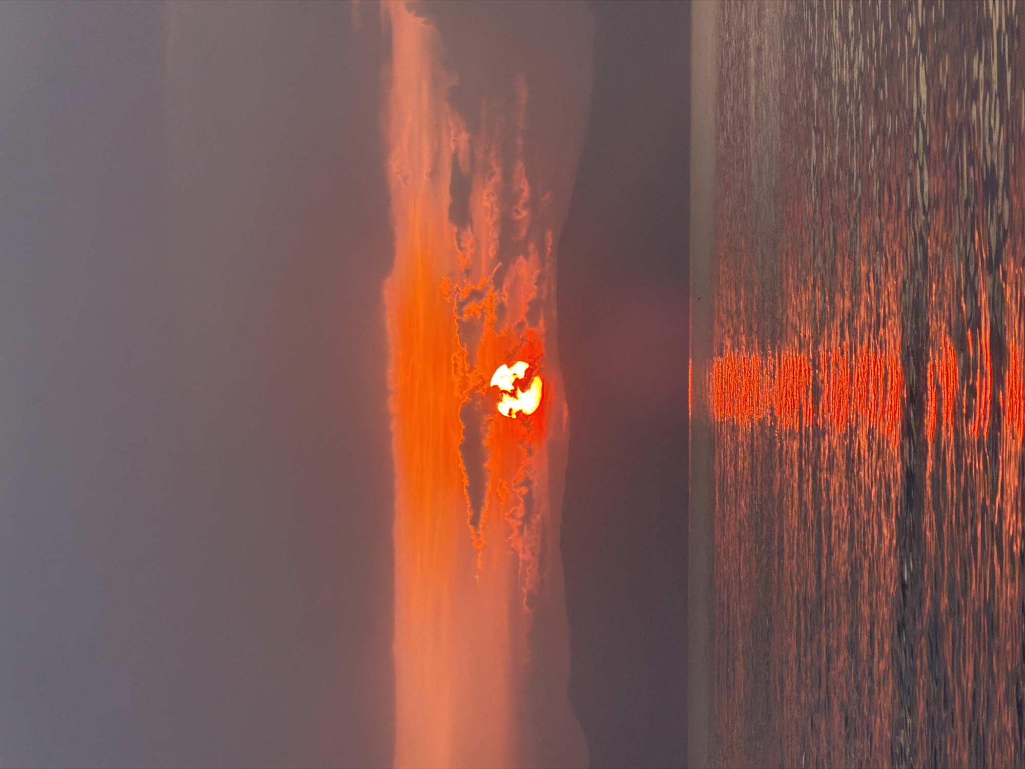 The image captures a sunset over a body of water, likely the ocean, near the city of Dong Ninh Hoa Ward, Vietnam. The sun, a vibrant orange orb, is partially obscured by dark, wispy clouds, casting a fiery glow across the sky and reflecting intensely on the rippling surface of the water. Silhouetted mountains form a dark, hazy line on the horizon between the sky and the water. The overall color palette is dominated by warm oranges, reds, and purples, characteristic of a sunset. There are no people or specific objects visible that would identify a precise location within Dong Ninh Hoa Ward. The scene is a natural landscape dominated by atmospheric and aquatic elements.