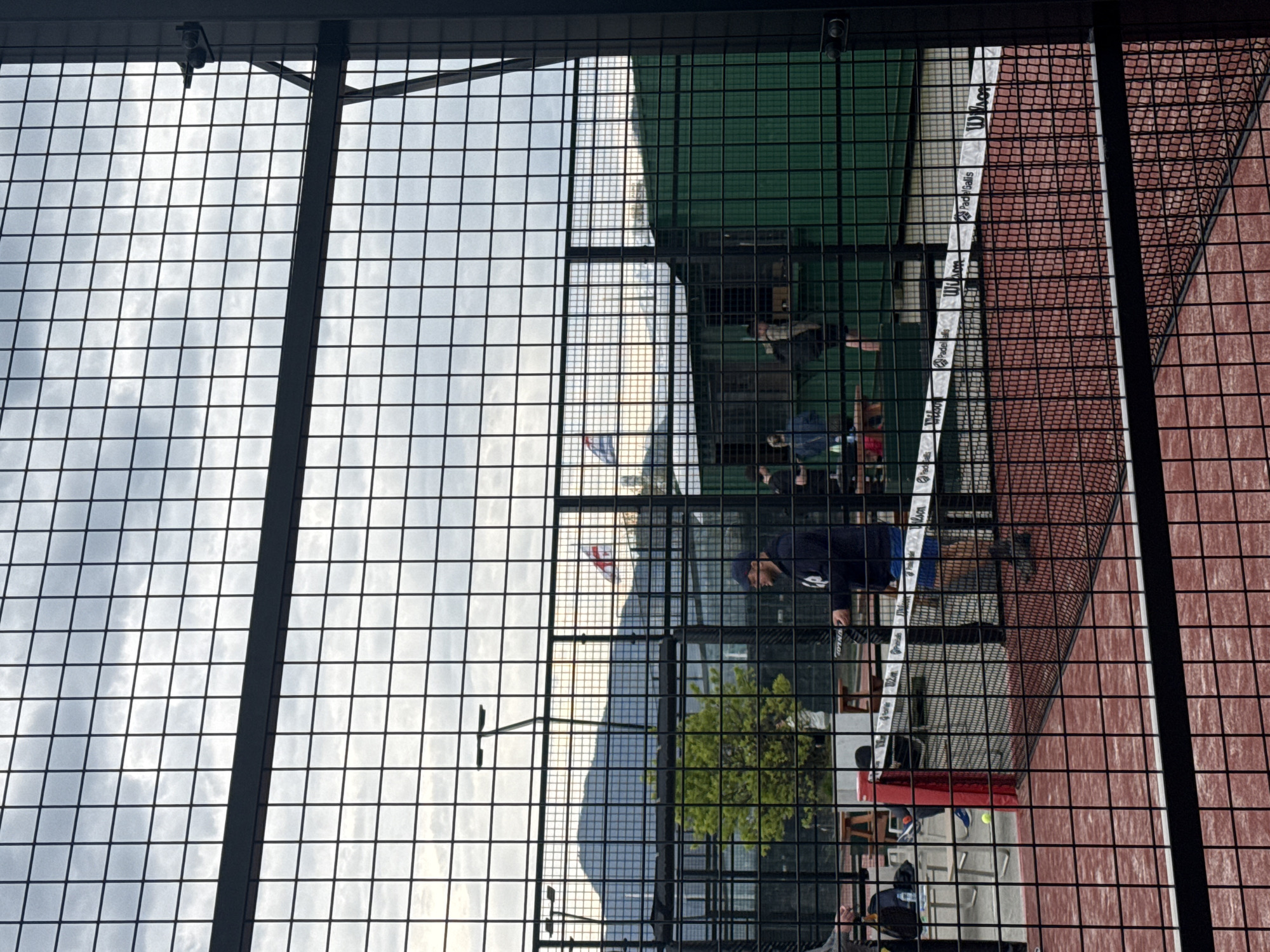 The image shows a tennis/paddle court viewed through a metal fence. The court is in Batumi, Georgia, as a Georgian flag can be seen in the background. Several people are gathered on and around the court, seemingly engaging in a game or practice session. The court has a green area and a red brick area separated by a net with the text "Wilson" on the net's fabric. There's also a mountain in the background and a cloudy sky.