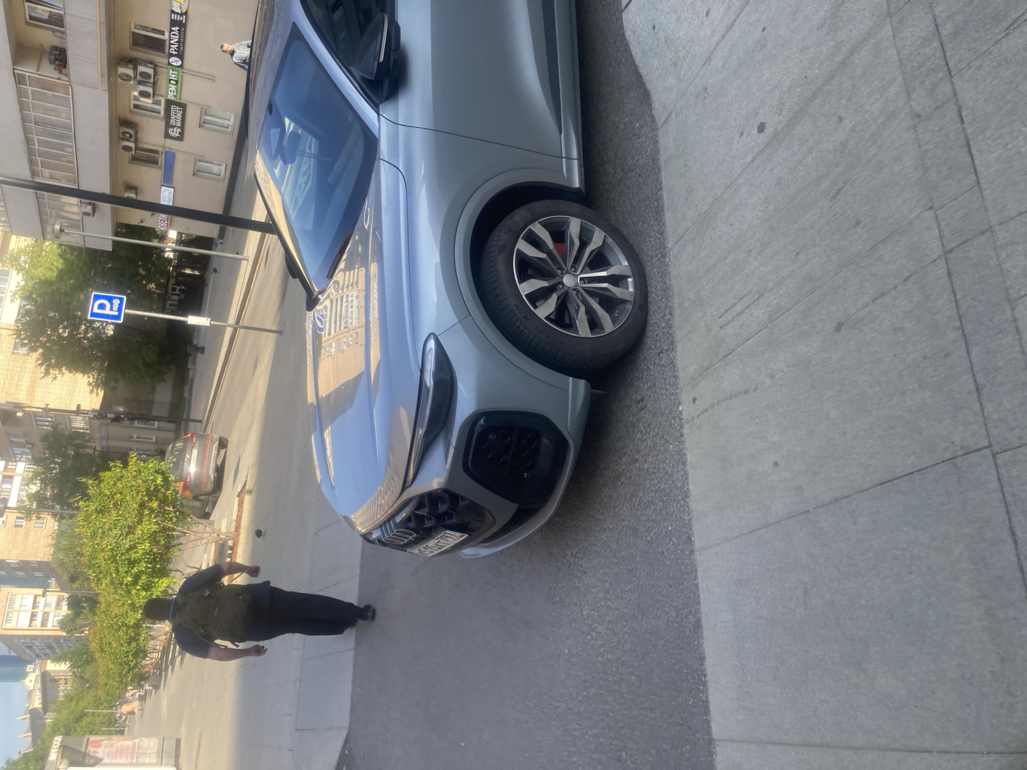 A horizontally-oriented image shows a silver Audi Q8 parked on the side of a street in Moscow, Russia. A man wearing a dark shirt and pants and carrying a backpack walks away from the car. The front of the car is in view, and the license plate, reading "1515AX797" is visible. In the background, there is a row of buildings, trees, a street with other cars parked on it, and a road sign. The ground is paved, and the lighting suggests it is daytime.
