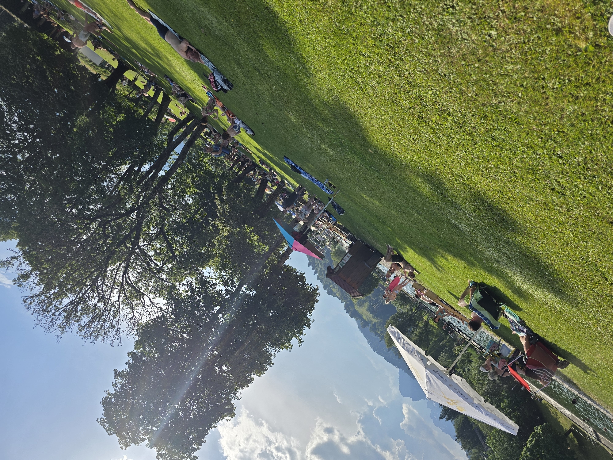 This is a tilted, wide shot of a grassy area next to a body of water, likely a lake or a pool, during a sunny day. The scene appears to be a recreational area with many people relaxing and enjoying the outdoors. The main subjects are groups of people scattered across the green lawn, some lying down, others sitting, and some are standing. They are dressed in casual summer attire, including swimwear.
Among the people, there are several notable objects. A prominent feature is a white hang glider with some text on its wing, positioned in the foreground, partially obscuring the view of the water and the people. Further back, there are colorful beach towels, bags, and what appears to be a small wooden structure with a roof, possibly a changing room or a kiosk. There are also some inflatable objects, one of which is bright pink and blue, adding to the playful atmosphere. The background shows a line of trees on the left side, with a clear blue sky and scattered white clouds above. Beyond the trees and the grassy area, rolling hills covered in greenery and mountains are visible, suggesting a scenic, possibly mountainous, location.
The activity taking place seems to be a leisurely day out, possibly a picnic or a gathering at a waterside park. The overall mood is relaxed and cheerful, indicated by the bright sunlight and the presence of people enjoying their leisure time. The sun's rays are visible, creating lens flares, which, along with the bright green grass and blue sky, suggest it is daytime, likely late morning or afternoon. The presence of the hang glider might imply that this is an area known for recreational aviation or a place where such activities are offered.
There is some visible text on the wing of the hang glider, but it is difficult to read clearly due to the angle and resolution. The setting of Garmisch-Partenkirchen, Germany, places this scene in a beautiful alpine region known for its outdoor activities and natural scenery.