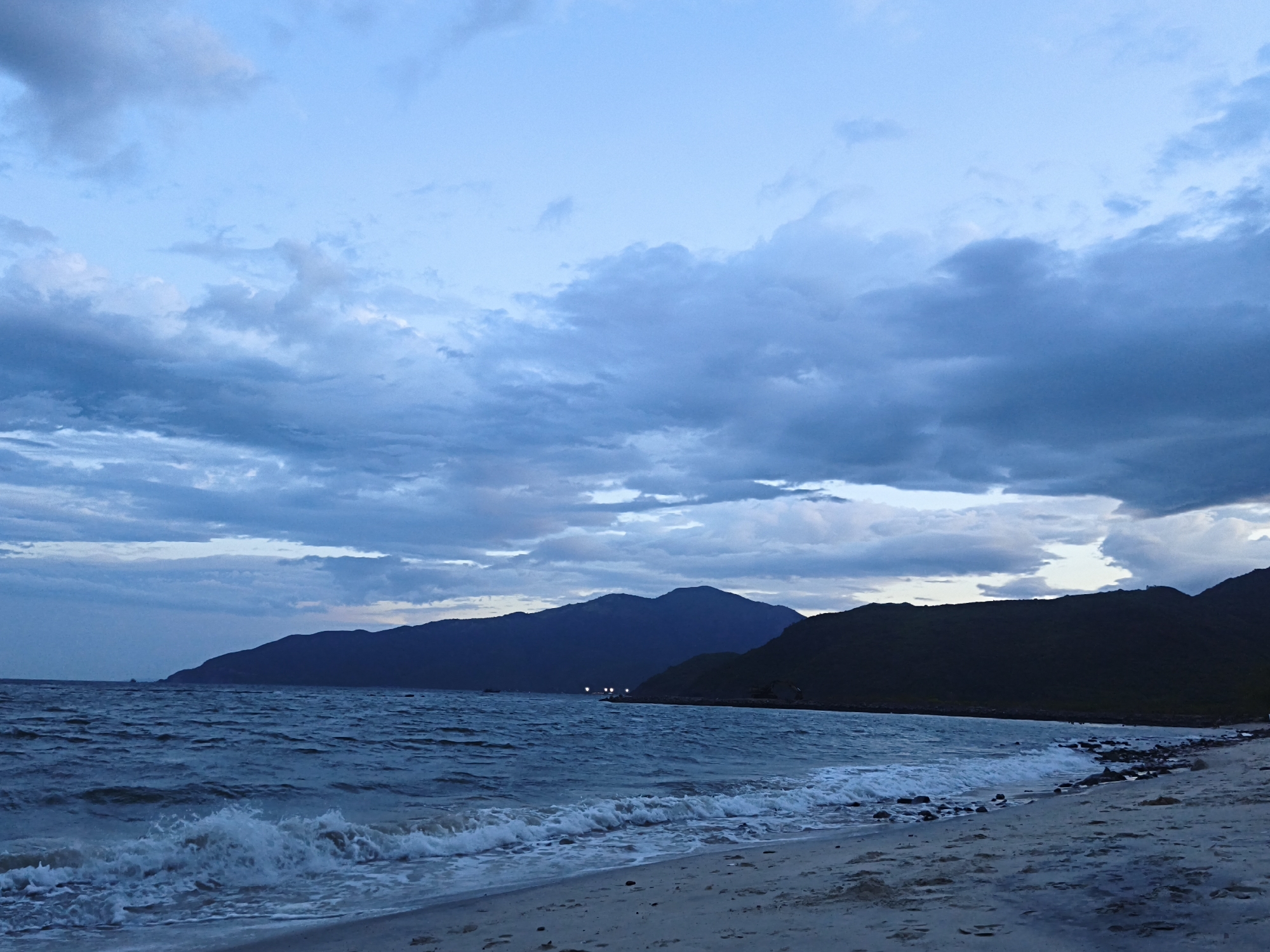 The image depicts a serene beach scene at dusk in Nha Trang, Vietnam. The sky is a soft blue with dramatic, dark clouds rolling in, hinting at approaching twilight. In the distance, silhouetted mountains form a majestic backdrop, their peaks softened by the fading light. The ocean is a deep blue, with gentle waves lapping at the shore, creating a foamy white edge along the sandy beach. The sand itself is a pale beige, showing faint footprints, suggesting recent human presence. A few scattered rocks are visible along the water's edge. The overall atmosphere is one of tranquility and natural beauty, captured in the soft, diffused light of the evening.