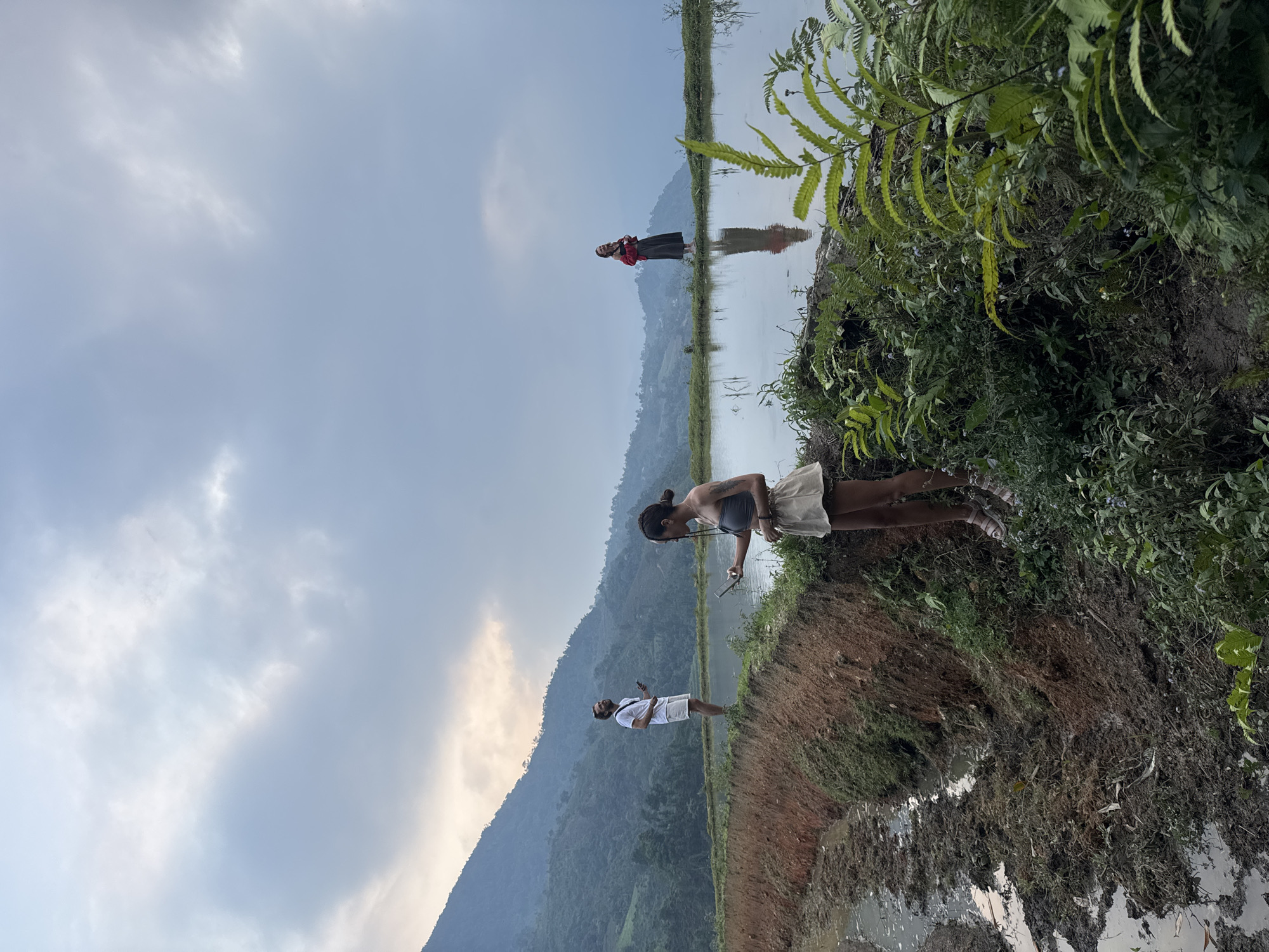The image captures a serene landscape in what appears to be a rural, mountainous region, consistent with Nậm Dịch Commune, Vietnam. The scene is dominated by vast, calm bodies of water, likely flooded rice paddies, which beautifully reflect the cloudy sky and distant mountains.
In the foreground, a woman with dark skin and dreadlocks, wearing a light crop top and skirt, stands on the edge of a muddy bank, looking down at an object in her hands, possibly a phone. Her lower legs and feet are visible. Adjacent to her, lush green ferns and other vegetation grow densely on the elevated ground.
Further back, on a narrow strip of land dividing sections of water, a man in a white t-shirt and light shorts stands, also appearing to look at a phone. His clear reflection is visible in the still water below him. Even further in the background, another individual dressed in dark clothing with a red top is visible on a similar strip of land, their reflection also distinct in the water.
The background features large, hazy mountains covered in dense green foliage, receding into the distance under a soft, overcast sky with diffused clouds. The overall lighting suggests either early morning or late afternoon, with no harsh shadows. The tranquil water, the reflective surfaces, and the distant mountains create a peaceful and scenic atmosphere. There is no visible text in the image.