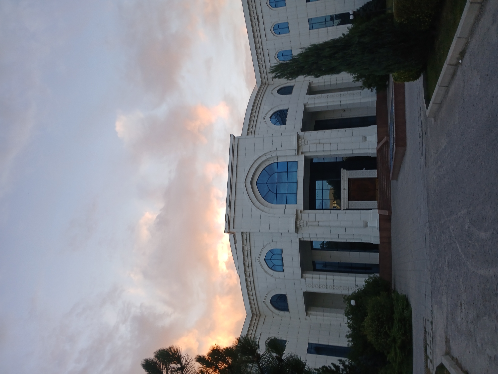 The image captures the exterior of a large, white, neoclassical building with arched windows and prominent columns. The building appears to be a government or public institution. The sky above is filled with dramatic, colorful clouds tinged with pink and orange, indicating either sunrise or sunset. In the foreground, there is a paved pathway leading to the building, with some greenery and trees on either side. The overall impression is one of grandeur and solemnity, set against a vibrant sky. Based on the architectural style and the mention of Tashkent, Uzbekistan, this building is likely the National University of Uzbekistan.