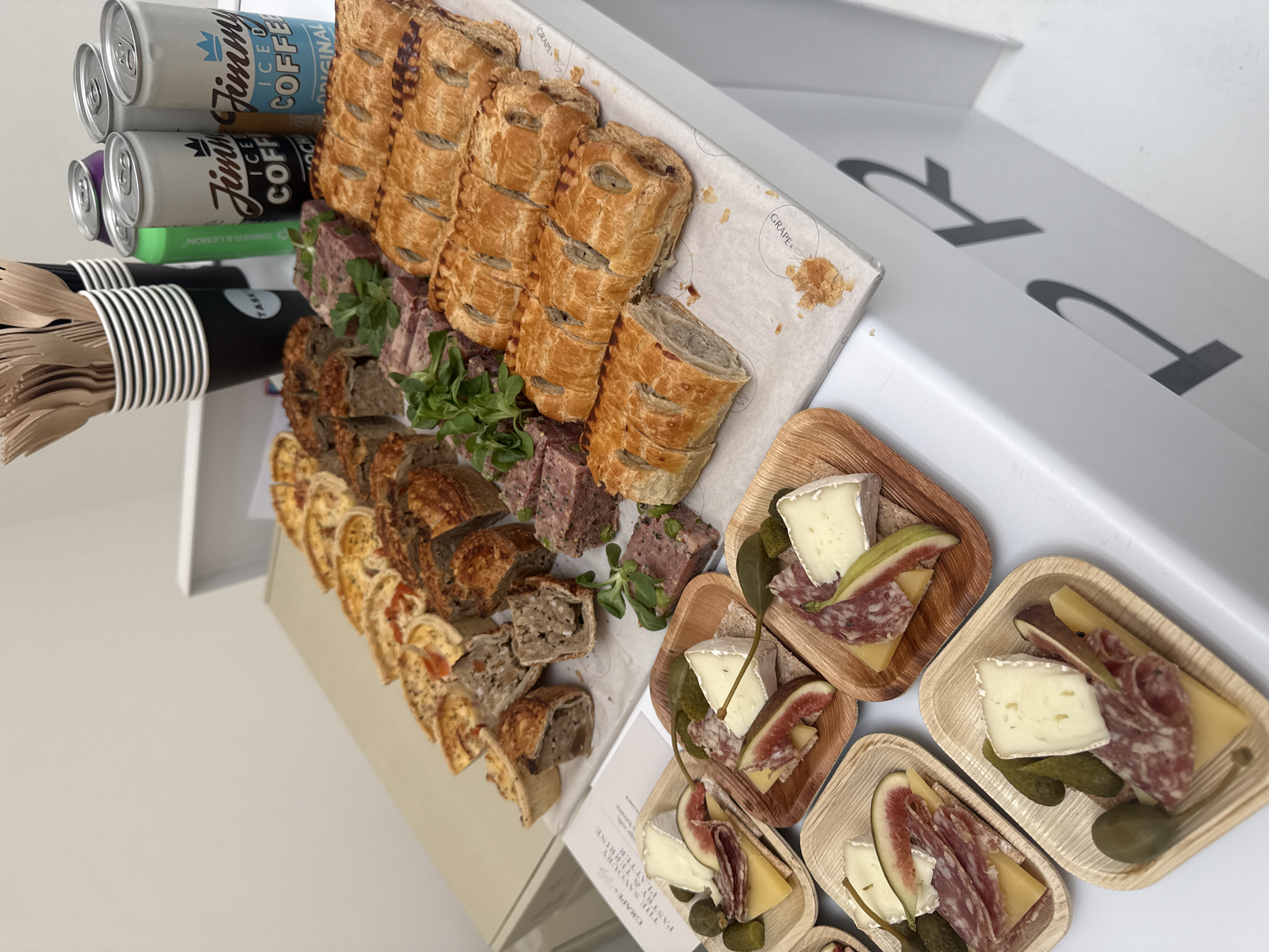 Here's a detailed description of the image:
**Scene:** An overhead shot of a catered food arrangement on a white surface, likely a counter or table. The focus is on a selection of savory pastries and cheese platters.
**Food Items:**
* Rows of sliced sausage rolls and other savory pastry rolls (possibly terrine).
* Small, individual serving platters containing cheese (likely Brie), slices of salami, pickles, figs, and pieces of baguette.
**Beverages & Utensils:**
* A collection of canned iced coffee drinks (branded "Jimmy's Iced Coffee") in different flavors (Original, Ginger & Lemon).
* A container filled with wooden cutlery (knives, forks, and spoons) alongside the beverages.
**Environment:**
* The food is presented in a catering box, as indicated by the branding ("GRAPE &").
* The surface appears to be clean and well-lit.
* The background is primarily white, suggesting an indoor setting.
* There are a couple of "R" alphabets visible on the catering box.
**Overall Impression:** This scene suggests a professional catering setup for a corporate event or meeting. The food selection is diverse, with both savory pastries and gourmet cheese platters. The arrangement indicates attention to detail and a focus on providing a convenient and appealing food experience.