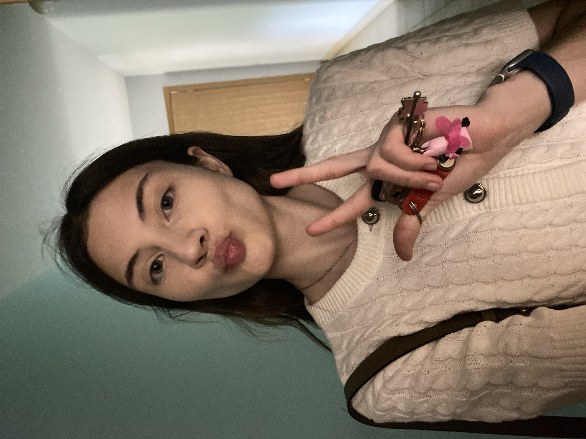 A young woman is taking a selfie in an indoor environment. She is oriented towards the camera, angled from the left. She has dark hair and fair skin. She is wearing a cream-colored cable-knit sweater and a blue smartwatch on her left wrist. Her left hand is raised, holding a set of keys with a pink charm, and she is making a peace sign with her fingers. She is making a kissing face expression. In the background, a light green wall is visible on the left, and a light-colored wooden door is seen partially in the center background. The exact location in Votkinsk, Russia, cannot be determined from this image alone.